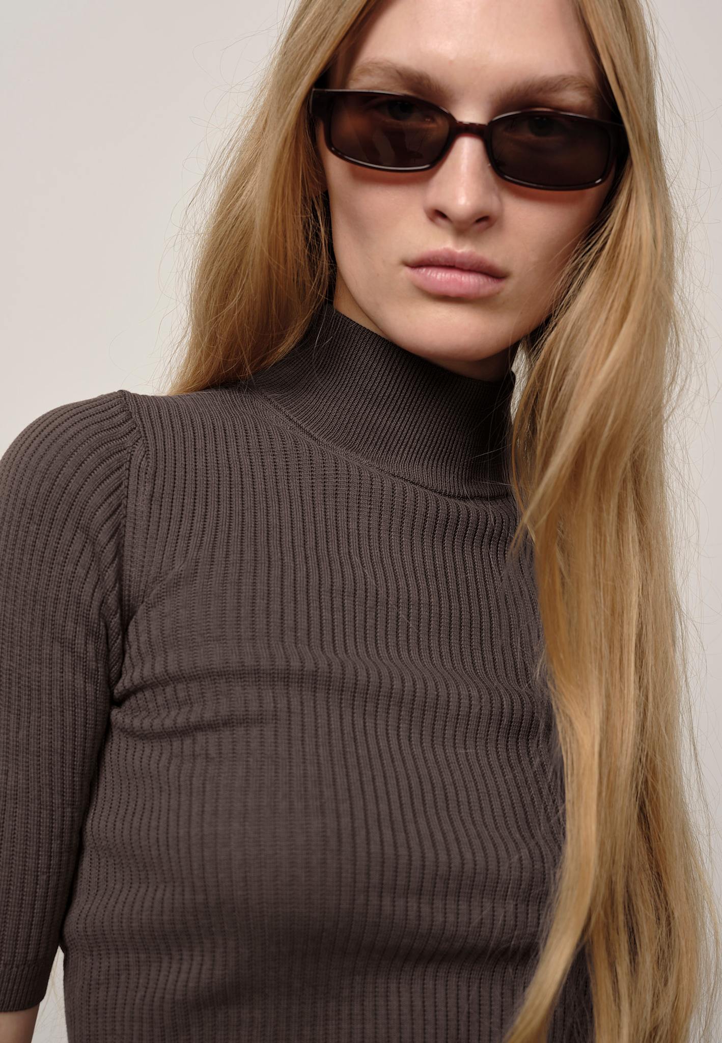 Person wearing a brown ribbed sweater and sunglasses with a plain background