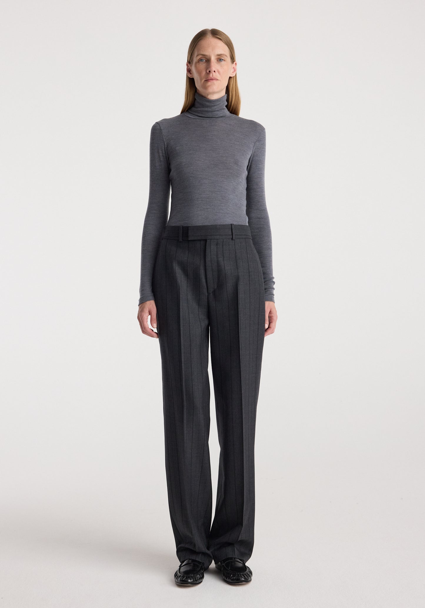 A model stands against a light gray studio background wearing a fitted gray mock-neck long-sleeve top and dark charcoal gray wide-leg trousers with a black belt.