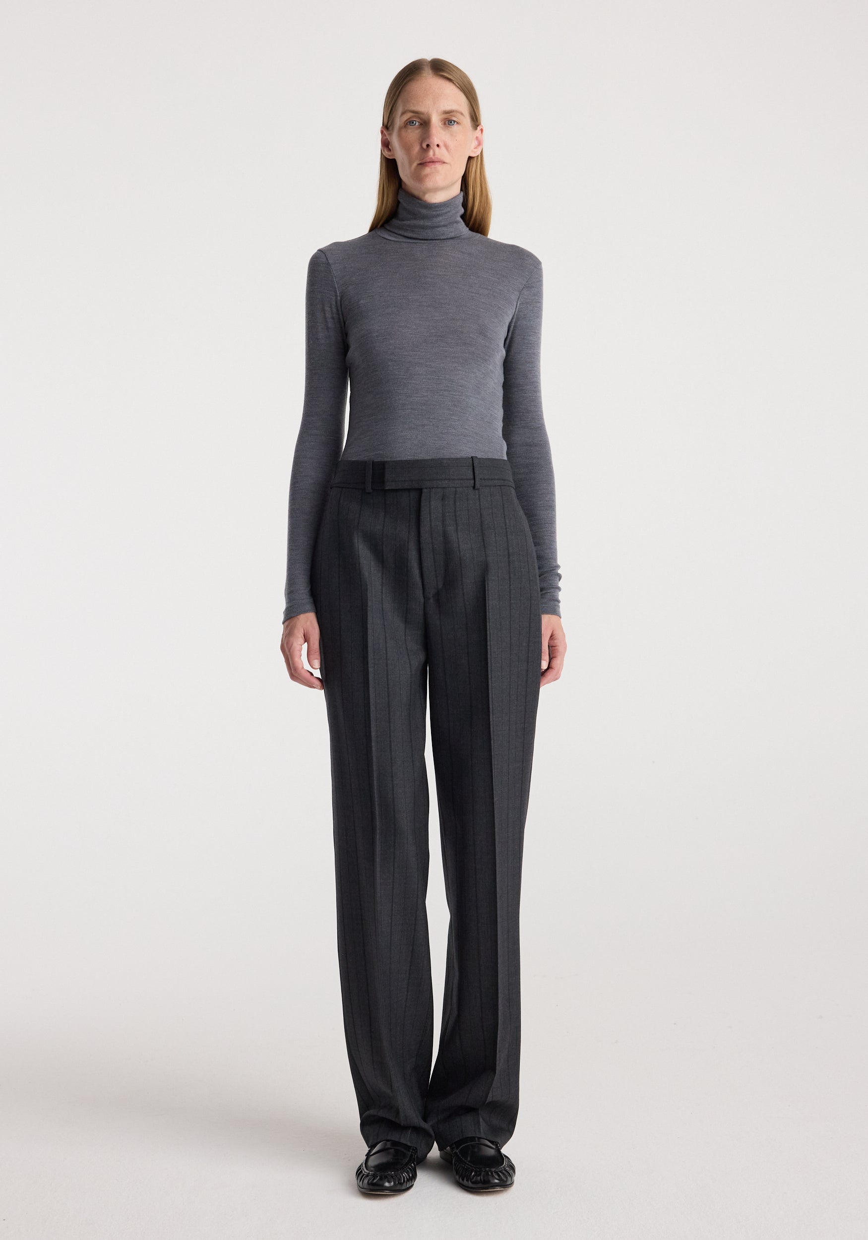 A model stands against a light gray studio background wearing a fitted gray mock-neck long-sleeve top and dark charcoal gray wide-leg trousers with a black belt.