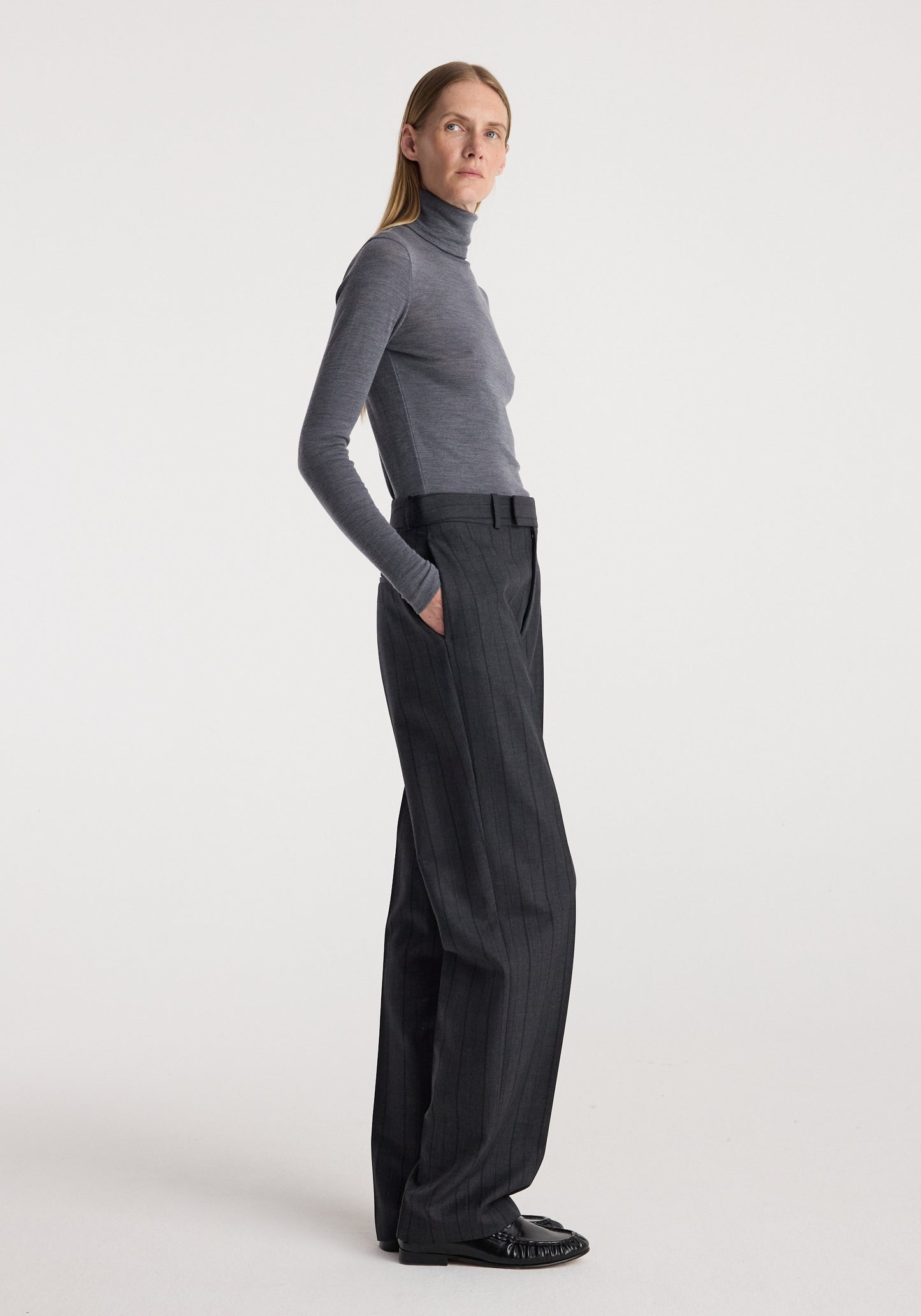 A woman stands in profile wearing a fitted gray turtleneck top and dark charcoal wide-leg trousers against a clean light gray studio background.