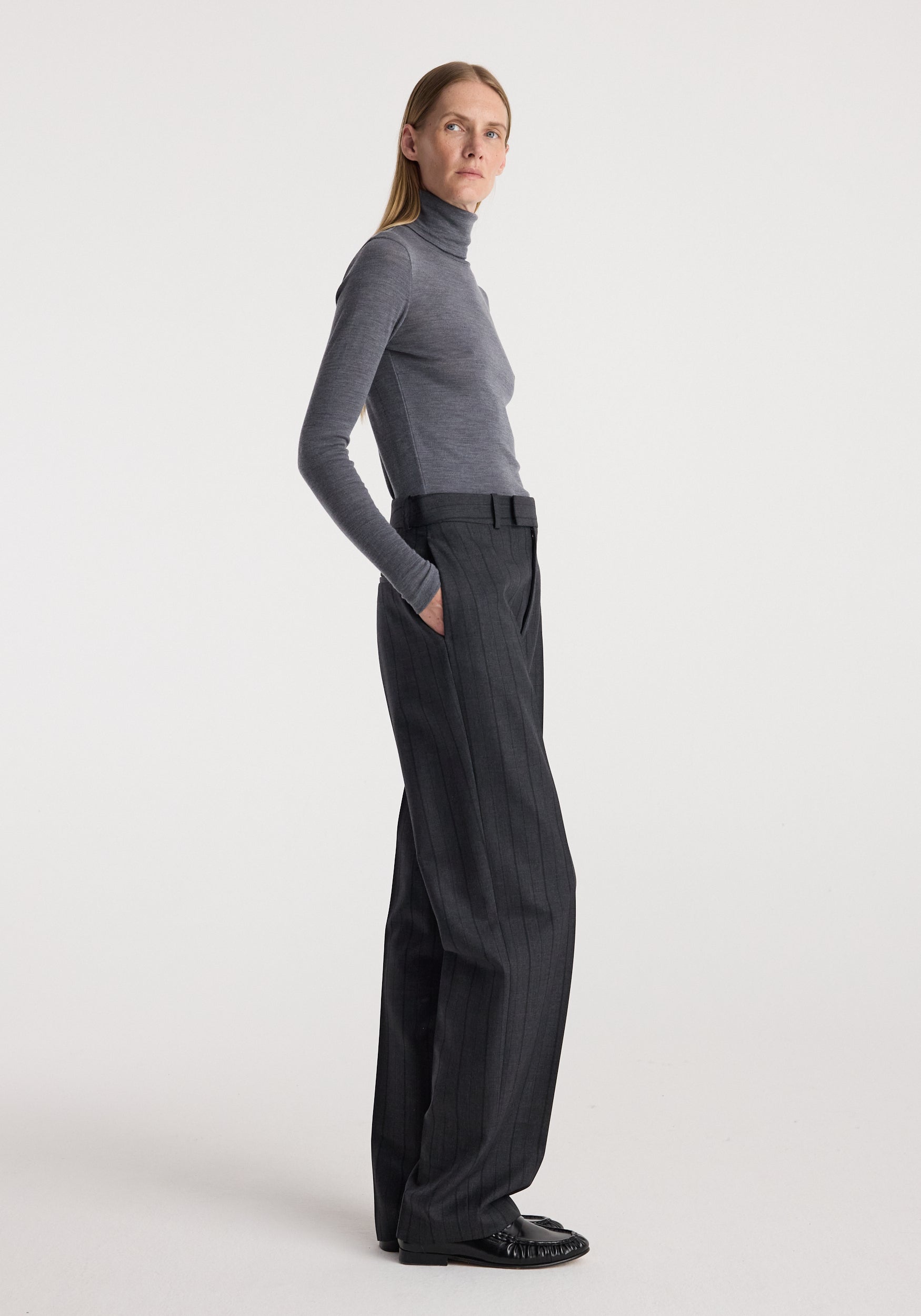 A woman stands in profile wearing a fitted gray turtleneck top and dark charcoal wide-leg trousers against a clean light gray studio background.