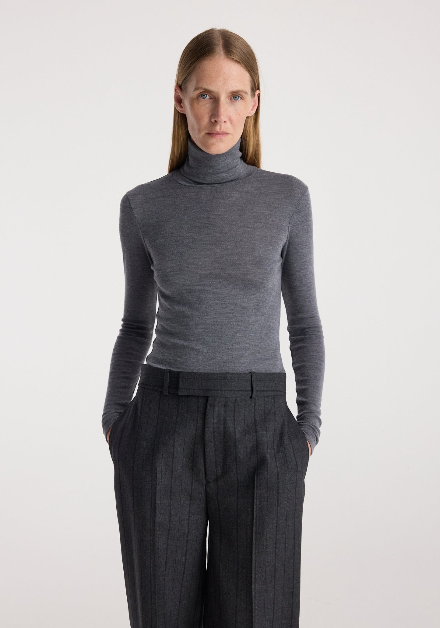 A woman modeling a fitted dark gray long-sleeve turtleneck sweater paired with dark charcoal pleated trousers, photographed against a clean light gray studio background.