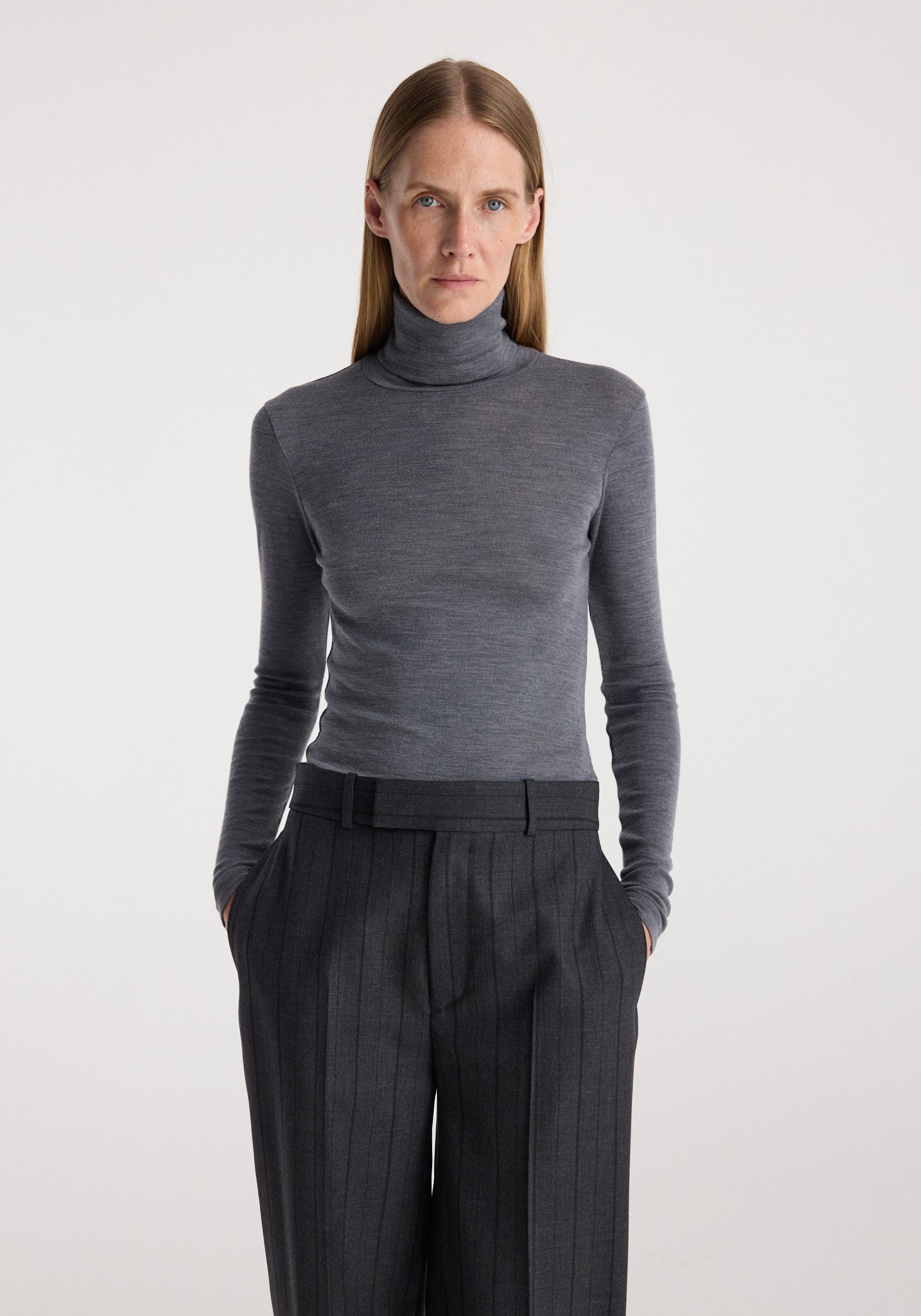 A woman modeling a fitted dark gray long-sleeve turtleneck sweater paired with dark charcoal pleated trousers, photographed against a clean light gray studio background.