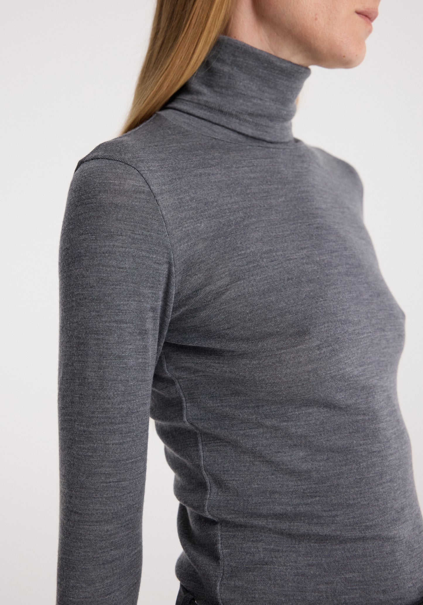 A close-up side view of a woman wearing a dark gray heathered turtleneck long-sleeve top against a plain light background.