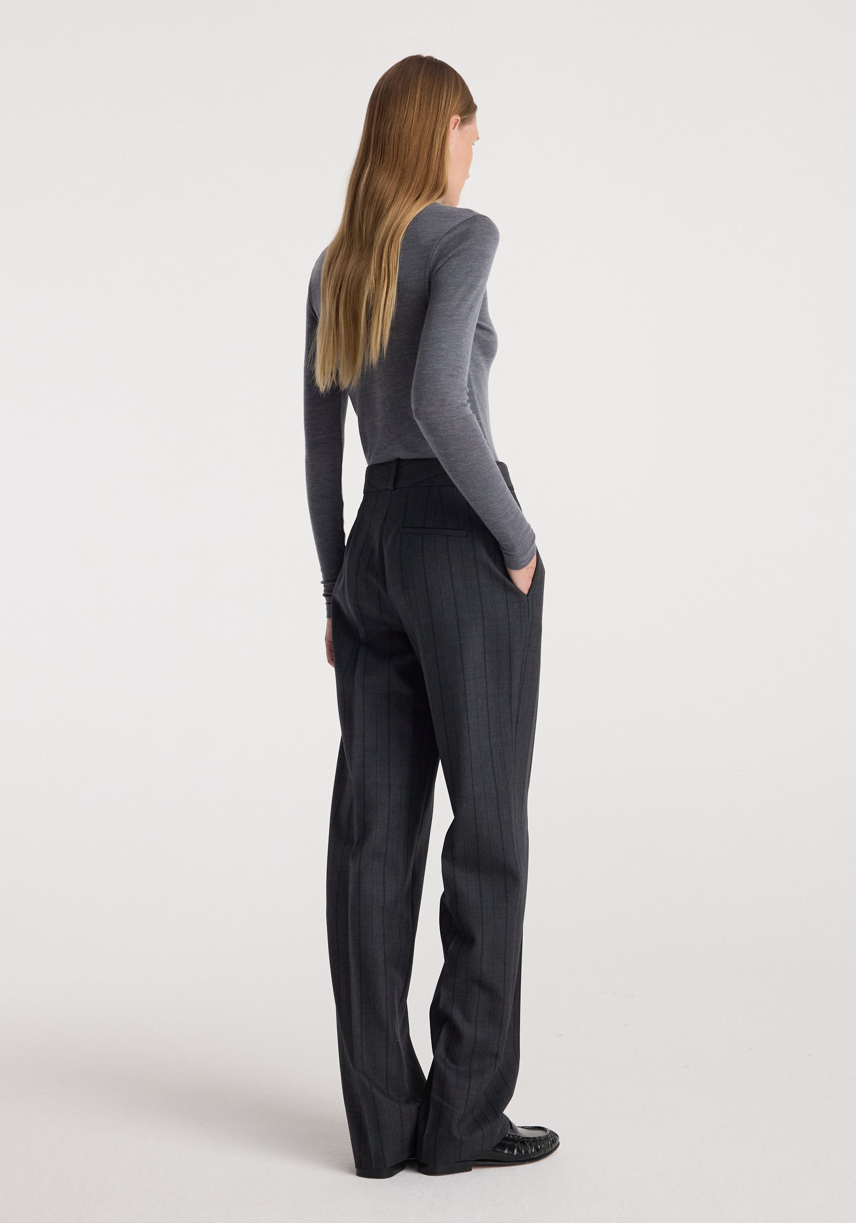 A woman is shown from the back wearing a gray long-sleeve top and dark charcoal gray pinstriped trousers, standing against a plain light gray studio background.
