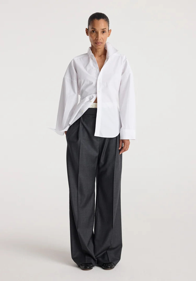 A model stands against a light gray studio background wearing an oversized white button-up shirt tucked into high-waisted, wide-leg charcoal gray trousers.