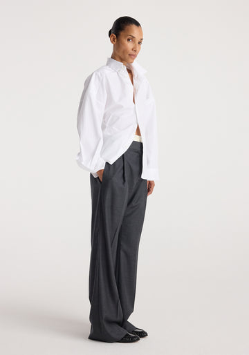 A model stands in profile wearing an oversized white button-down shirt, tucked into dark charcoal gray wide-leg trousers, against a plain light gray studio background.