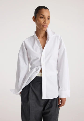 A model wearing a white long-sleeve button-down shirt with a relaxed, oversized fit and dark charcoal trousers, photographed against a neutral light gray studio background.