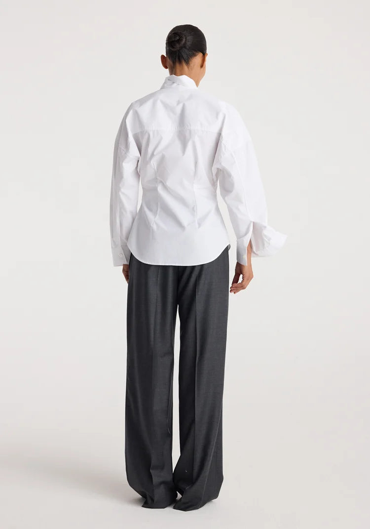 A model shown from the back wearing a white long-sleeve button-up shirt with dramatic ruffled cuffs and dark charcoal gray wide-leg trousers, set against a plain light gray studio background.