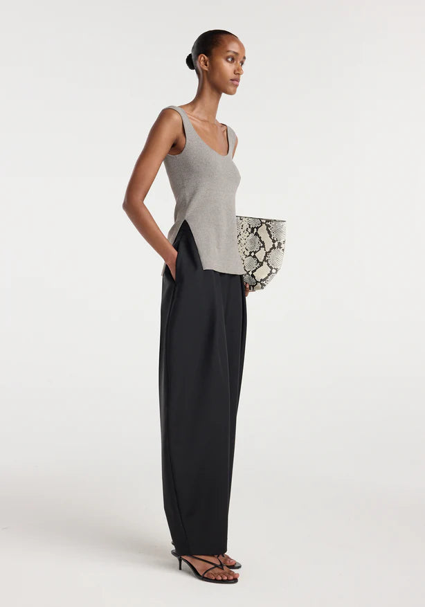 Woman wearing a gray sleeveless top and black pants, holding a patterned clutch against a white background