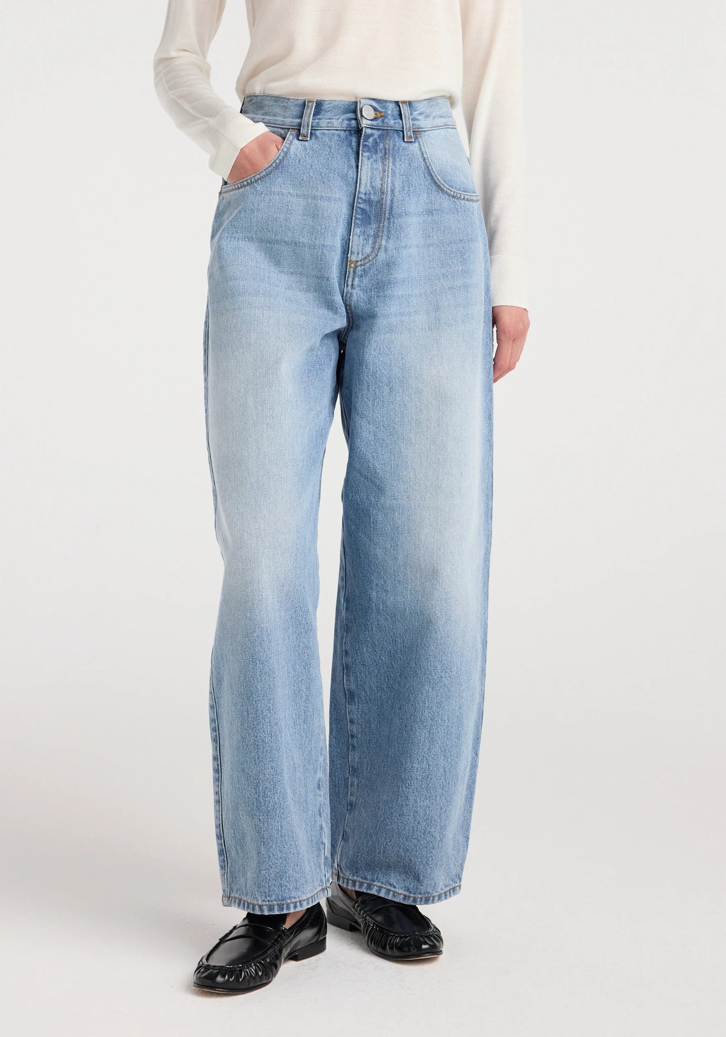 A person wearing light-wash blue wide-leg jeans with a cream long-sleeve top and black loafers, photographed against a plain light gray studio background.
