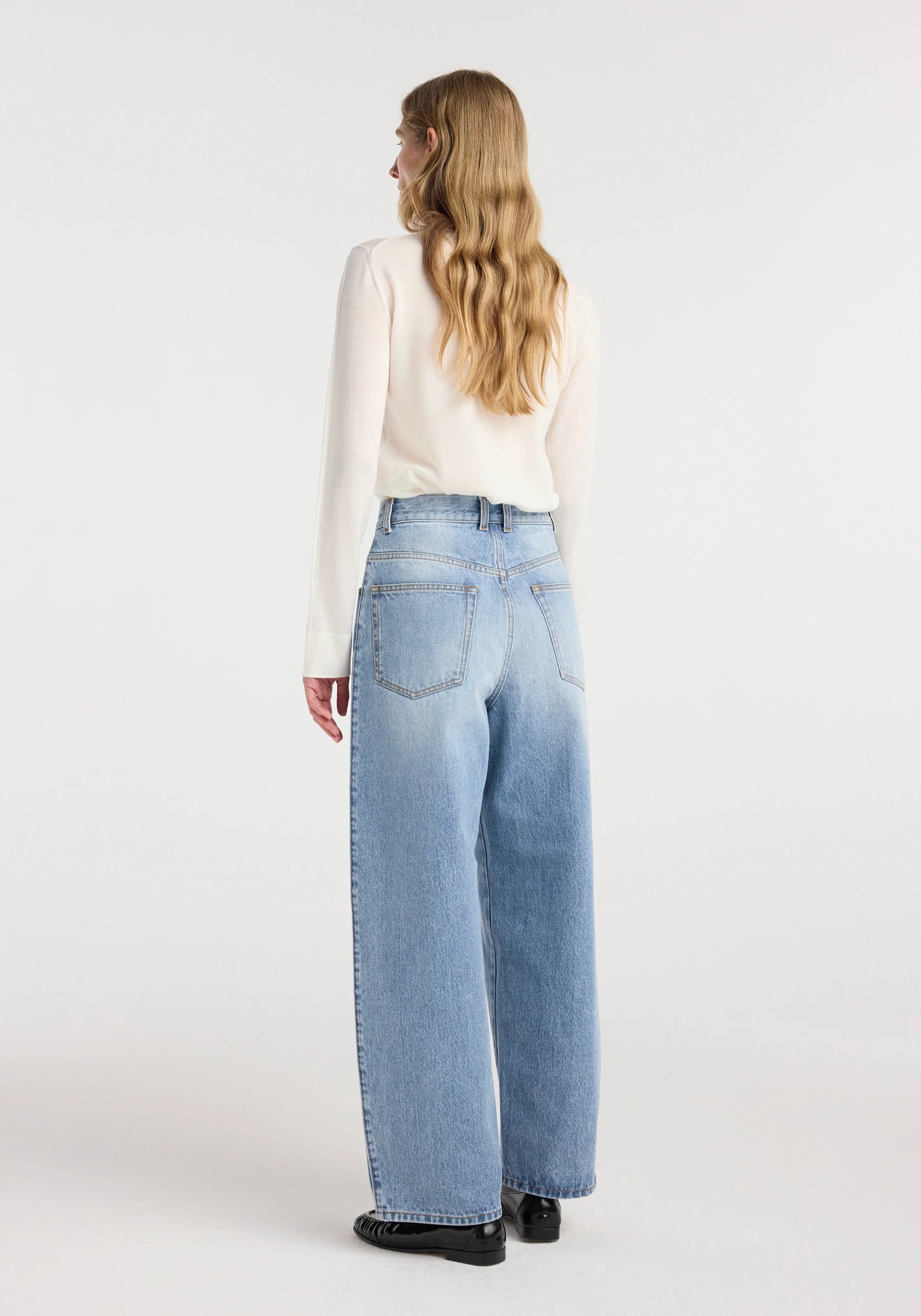 A woman shown from the back wearing light-wash wide-leg jeans, a cream long-sleeve top, and black shoes against a plain light gray studio background.