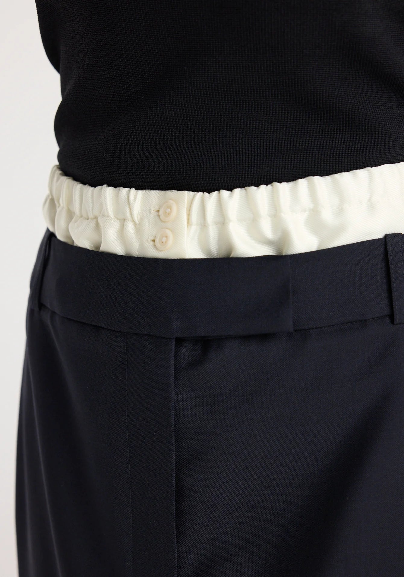 Close-up of a person wearing a black sweater with a white waistband and navy pants on a light background.