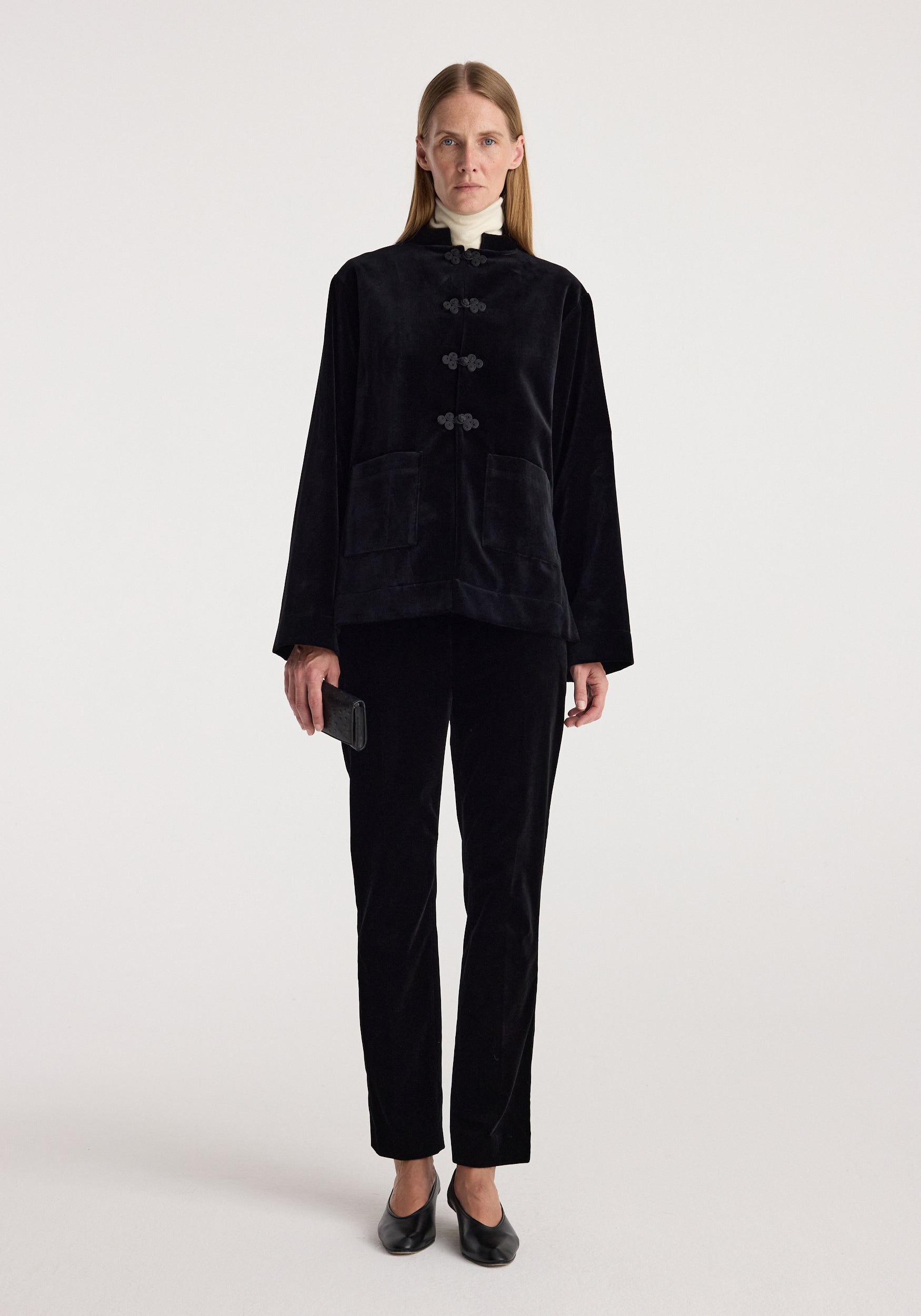 A model stands against a light gray studio background wearing a black velvet two-piece set consisting of a cropped jacket with toggle closures and matching trousers.