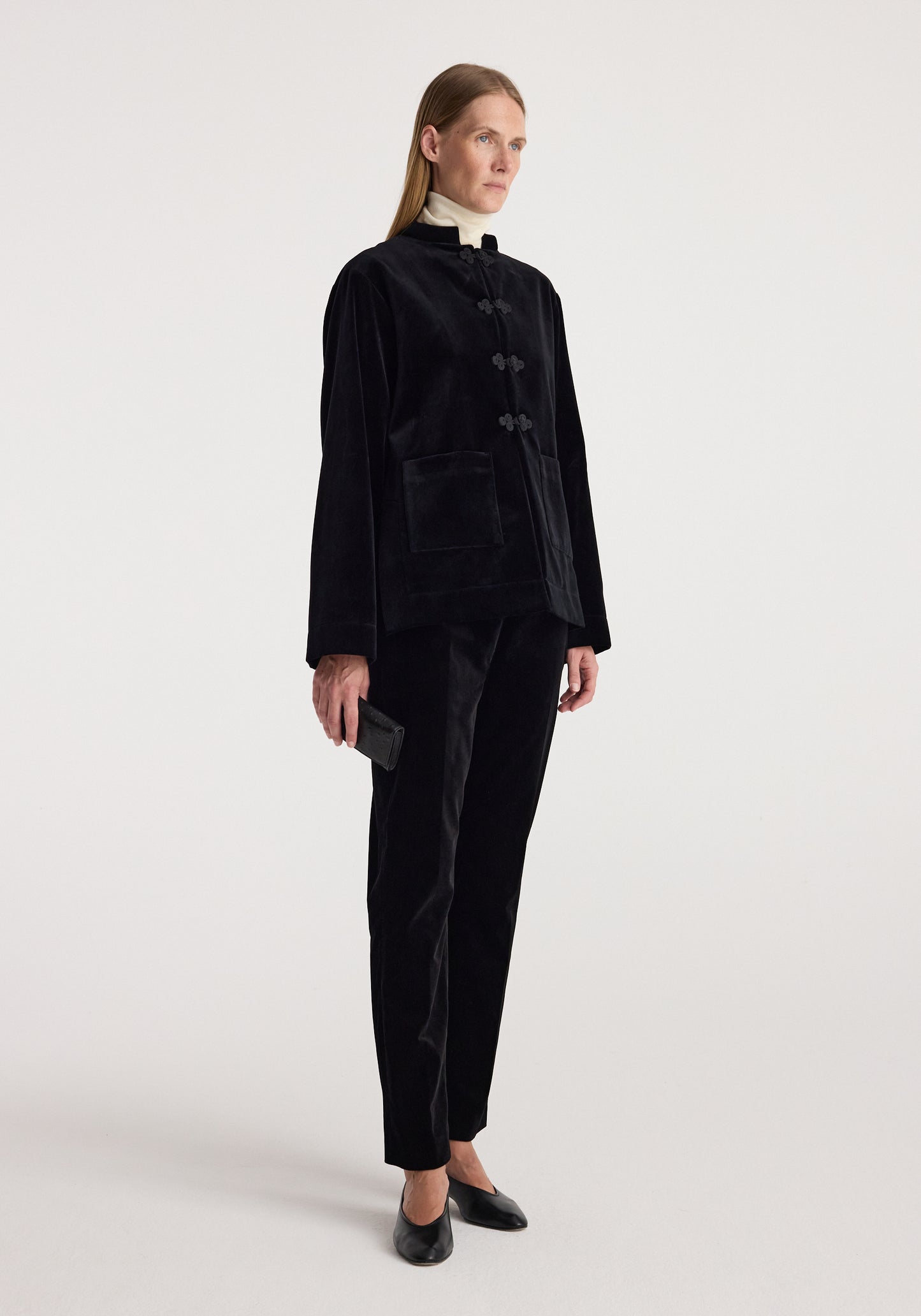 A model wearing a black velvet two-piece suit consisting of a long-sleeve jacket with traditional frog closures and matching trousers, paired with black leather shoes against a light gray studio background.