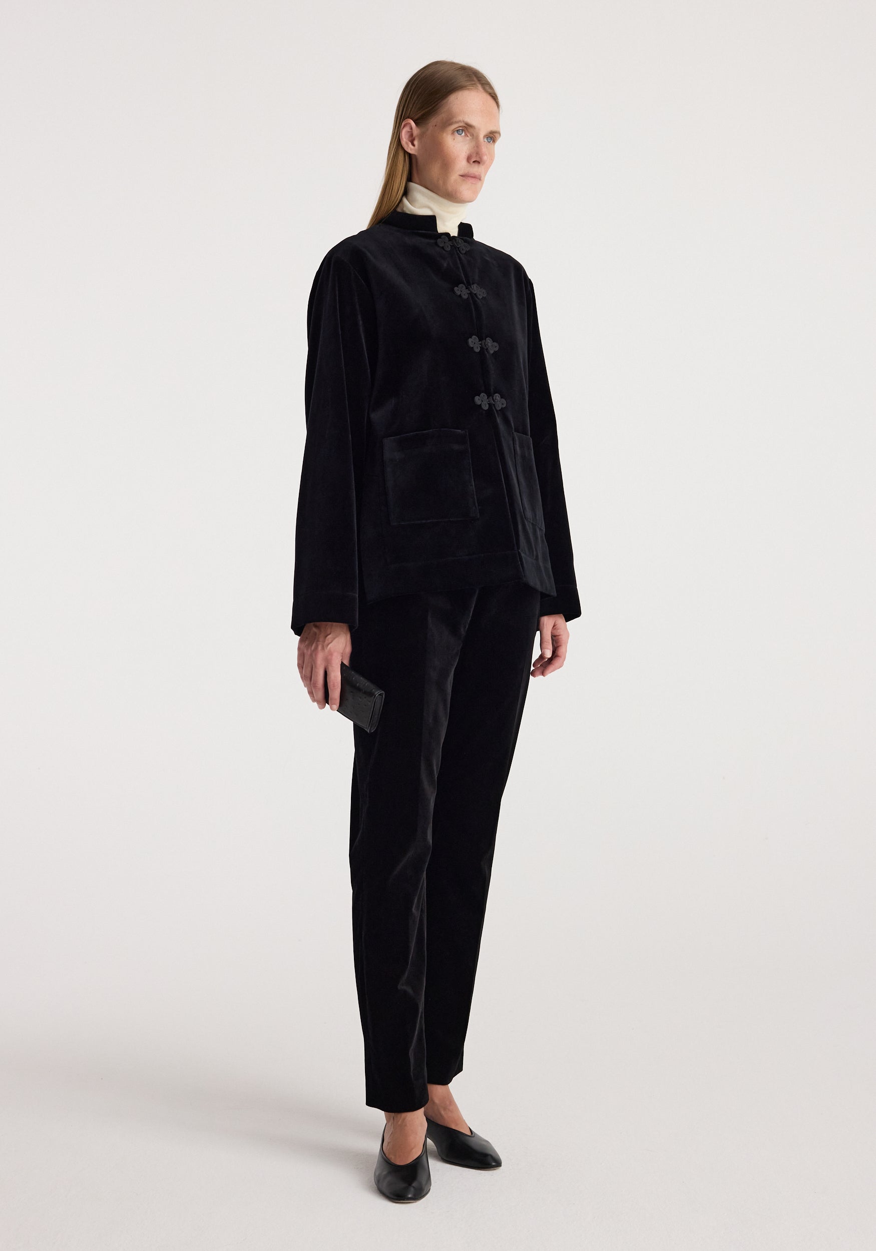 A model wearing a black velvet two-piece suit consisting of a long-sleeve jacket with traditional frog closures and matching trousers, paired with black leather shoes against a light gray studio background.