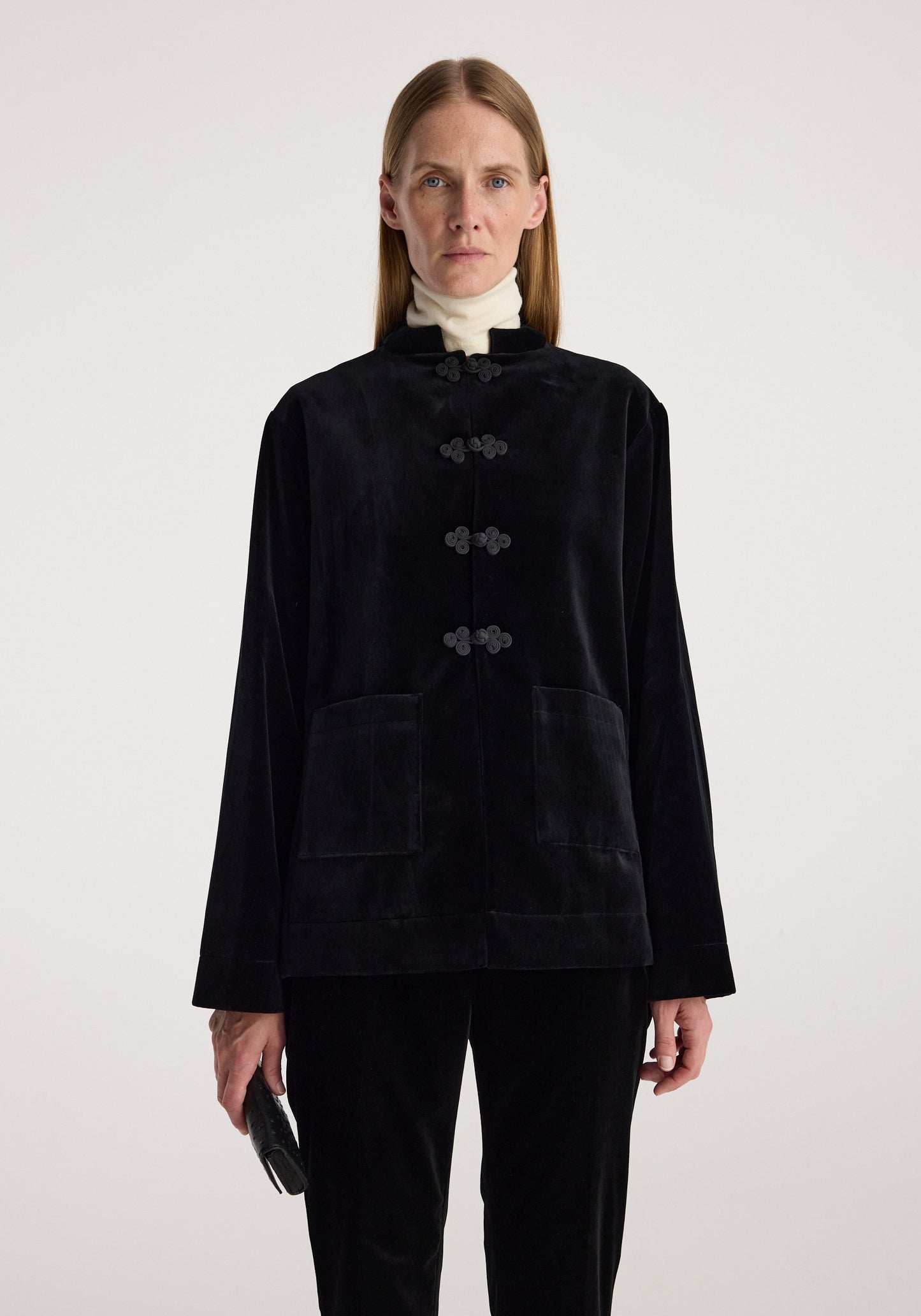 A model wearing a black velvet jacket with traditional Chinese-style frog closures and front patch pockets, paired with black trousers and a white turtleneck, against a plain light gray background.