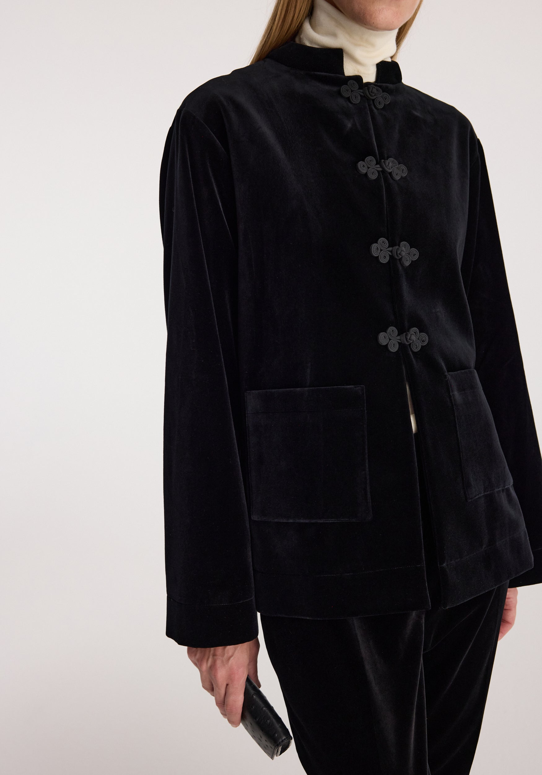 A person wearing a black velvet jacket with four decorative button closures on a stand collar, paired with black velvet pants and a cream turtleneck, against a light gray studio background.