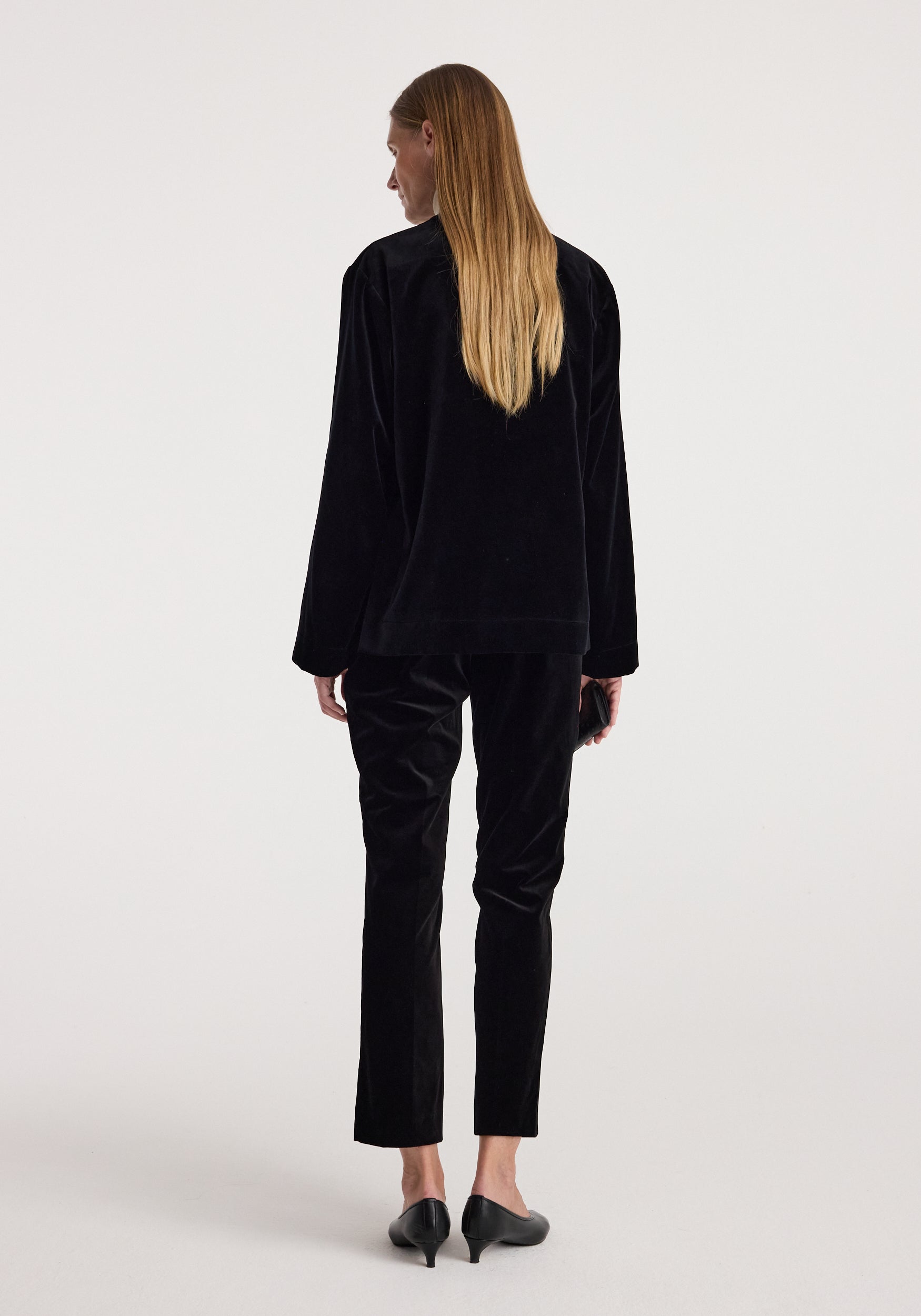 A woman with long blonde hair is shown from behind wearing a black velvet long-sleeve top and matching black velvet cropped trousers, paired with black pointed-toe heels, against a light gray studio background.