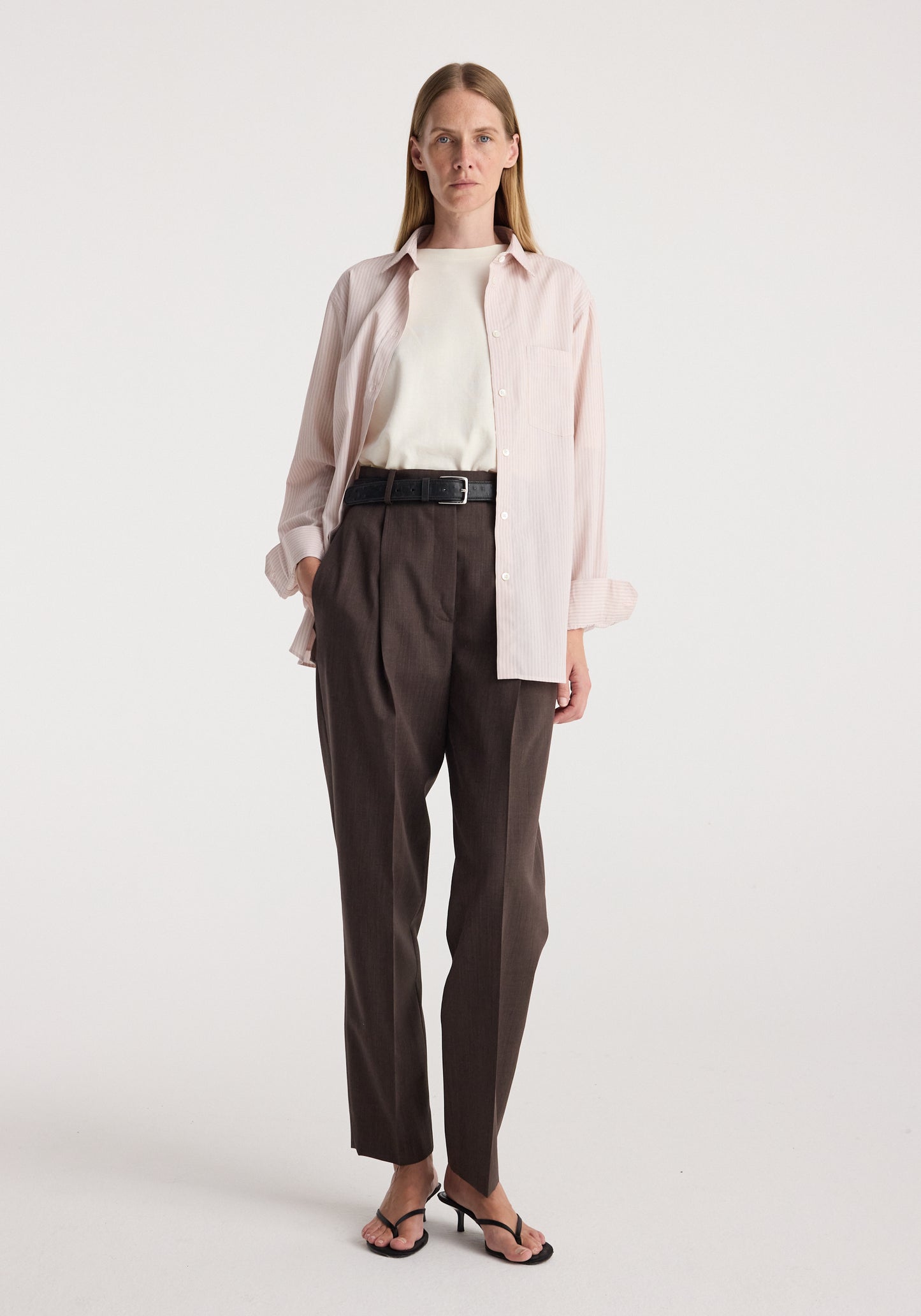 A woman stands against a plain light gray studio background wearing a light pink button-up shirt over a white top, dark brown pleated trousers with a black belt, and black sandals.
