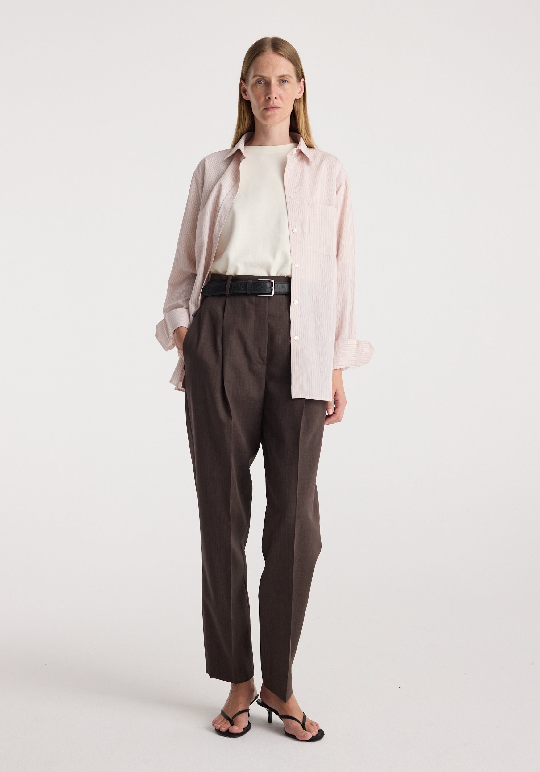 A woman stands against a plain light gray studio background wearing a light pink button-up shirt over a white top, dark brown pleated trousers with a black belt, and black sandals.