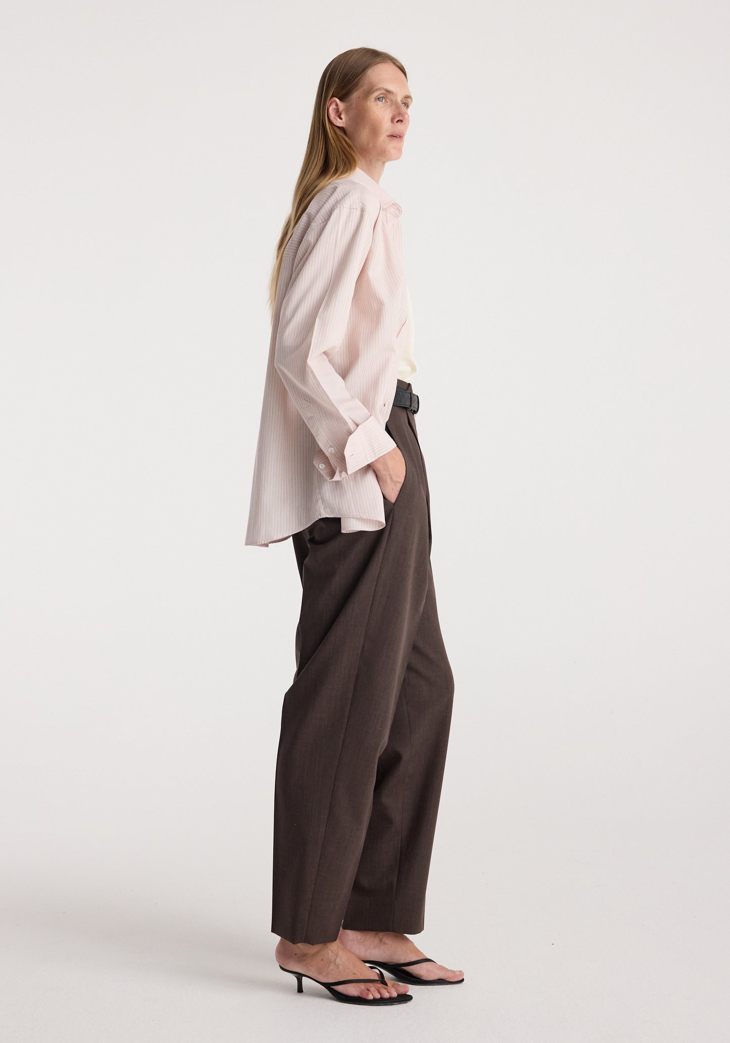 A woman stands in profile wearing a pale blush pink blouse with a ruffled collar and cuffs, dark brown wide-leg trousers, and black heeled sandals against a plain light gray studio background.