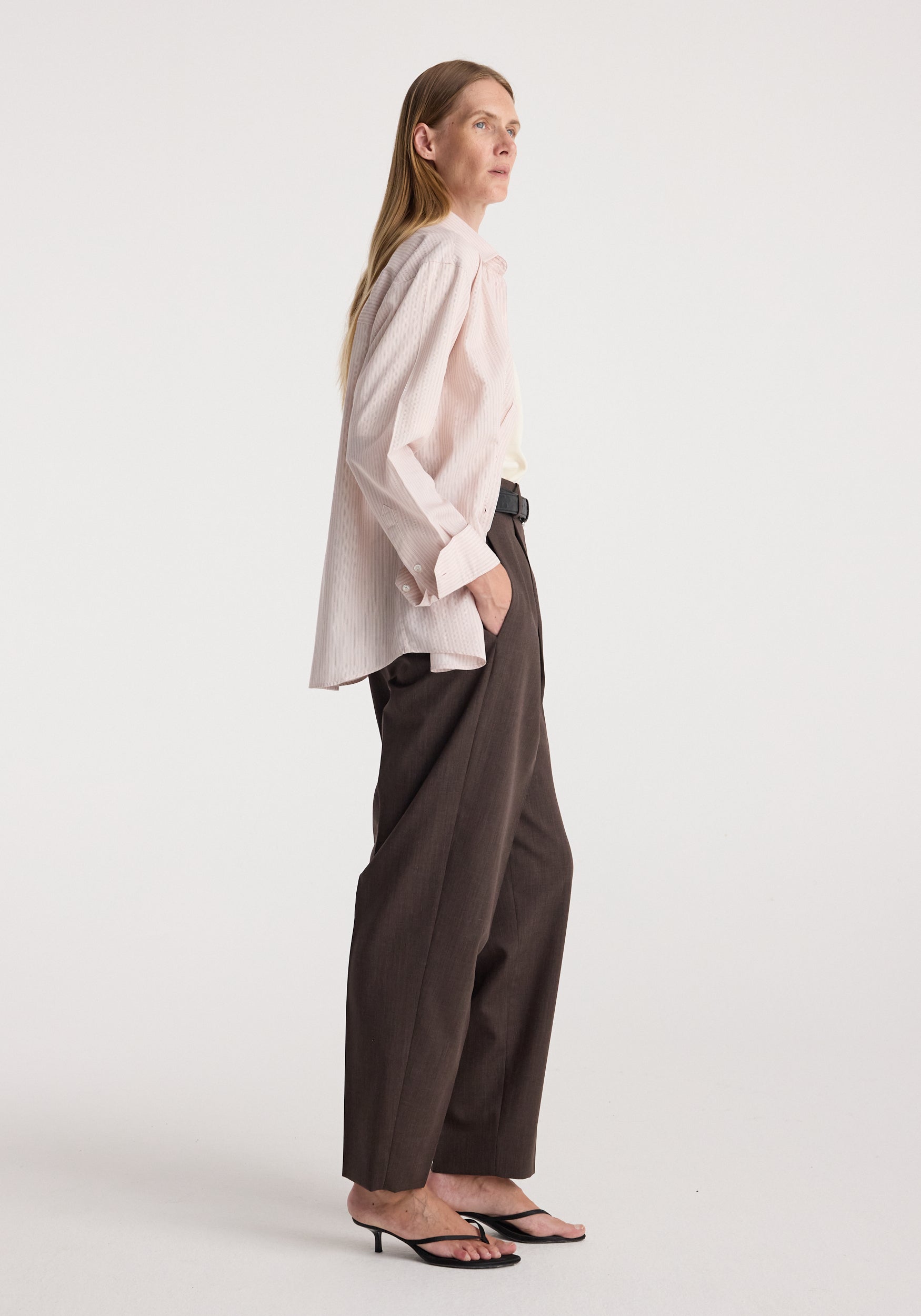 A woman stands in profile wearing a pale blush pink blouse with a ruffled collar and cuffs, dark brown wide-leg trousers, and black heeled sandals against a plain light gray studio background.