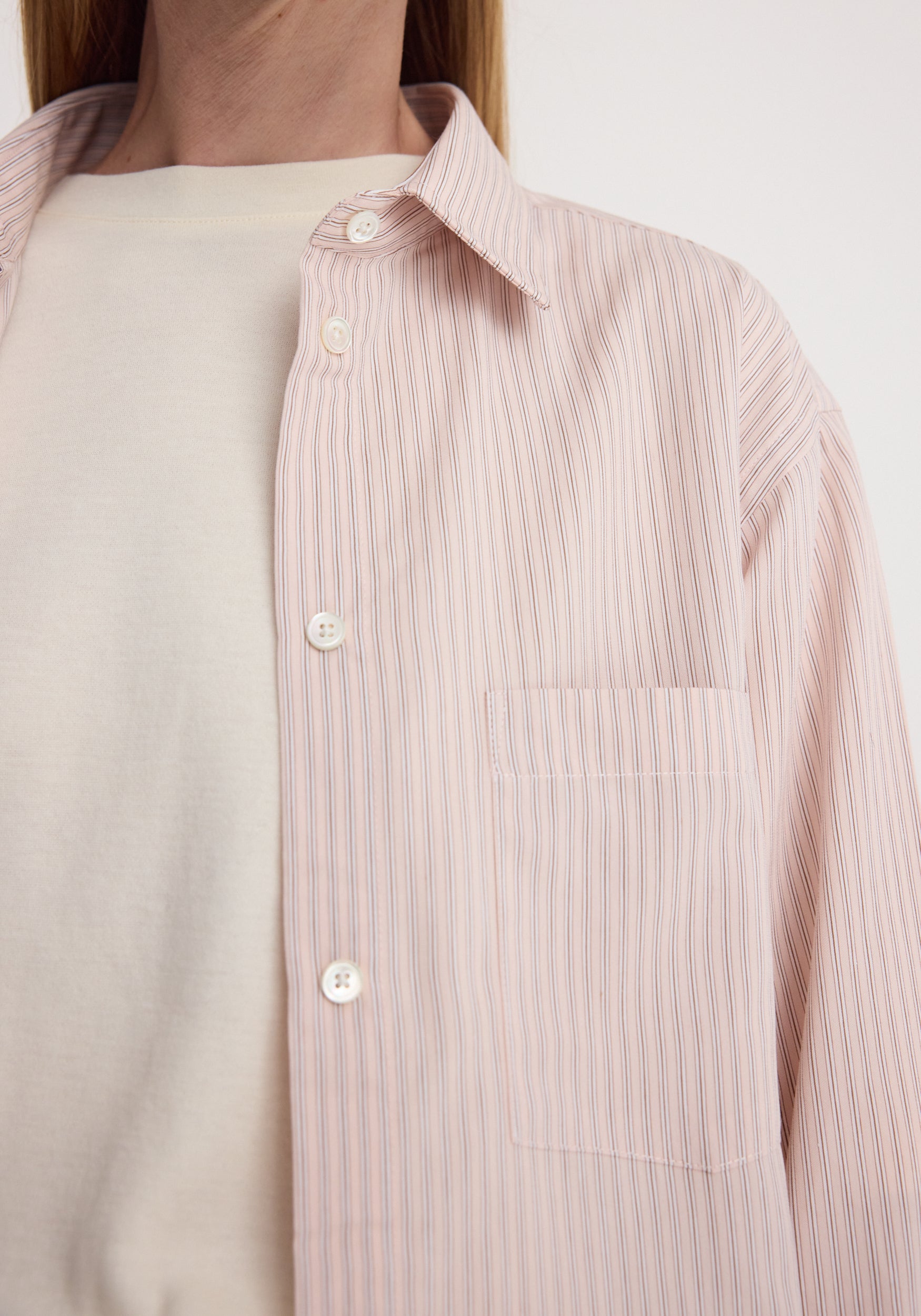 A close-up of a person wearing a light pink and white vertically striped button-down shirt over a cream-colored top.