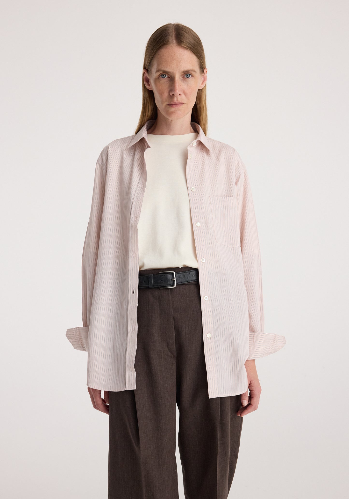 A model stands against a light gray studio background wearing a pale pink button-up shirt over a cream knit top, paired with dark brown trousers and a black belt.