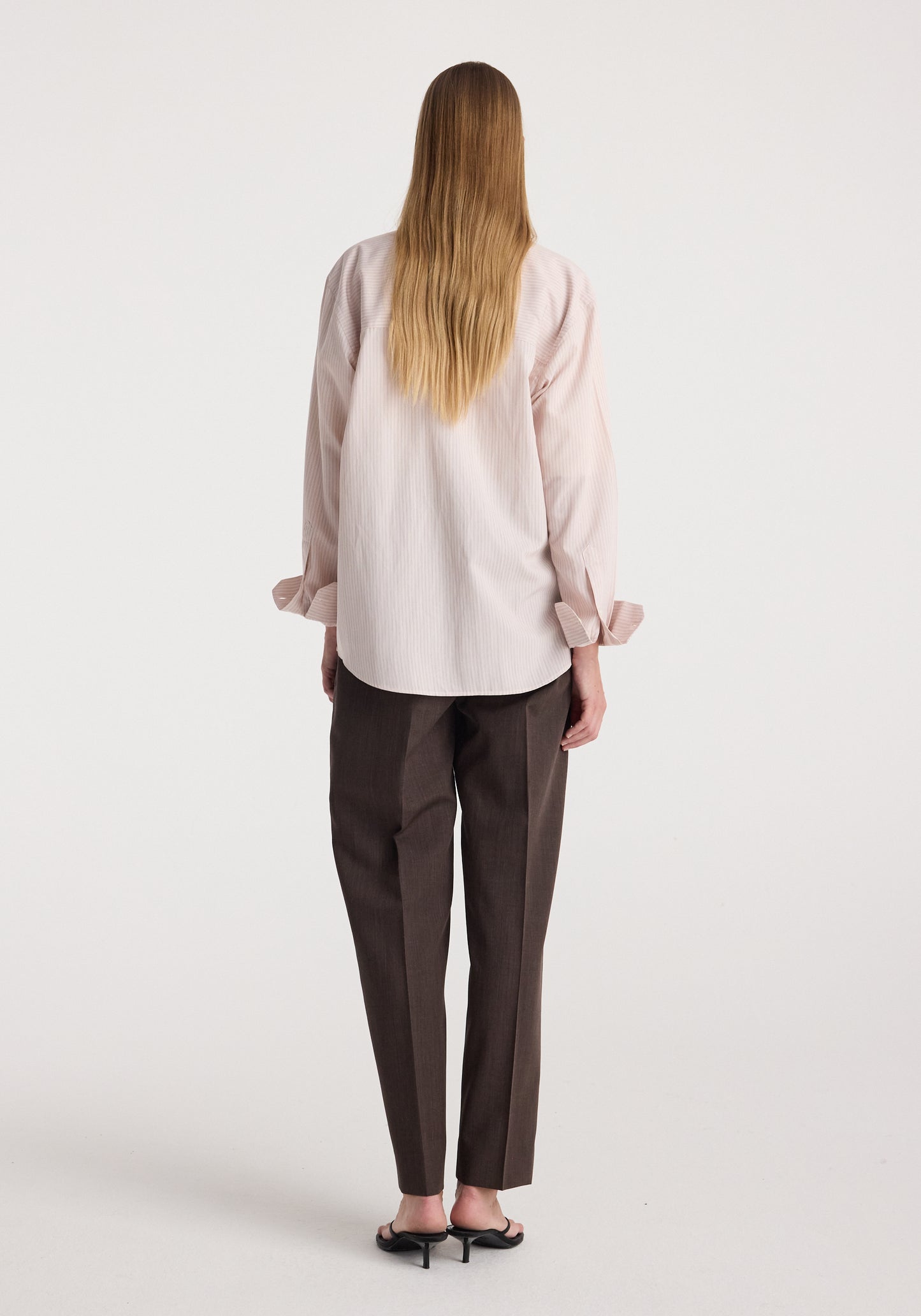 A woman viewed from behind wearing a light blush pink linen shirt with rolled cuffs and dark brown trousers, standing against a plain white studio background.