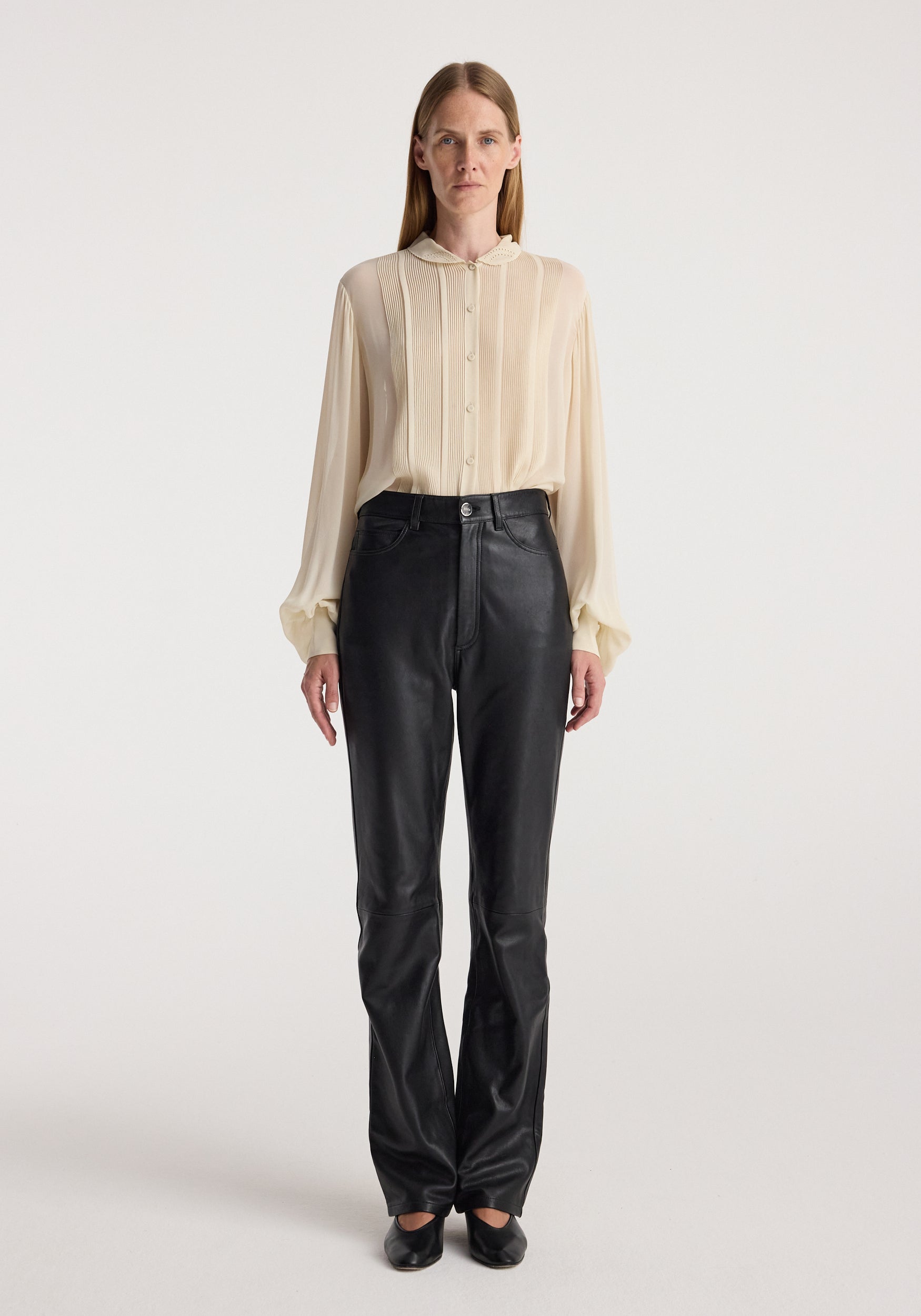 A model stands against a light gray studio background wearing a cream-colored sheer long-sleeve button-up blouse and black leather pants.