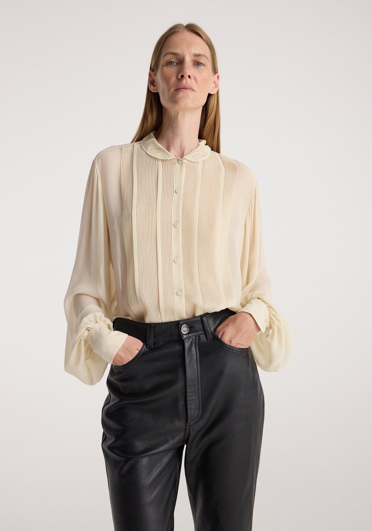 A woman modeling a cream-colored, semi-sheer long-sleeve blouse with pleated details and gathered cuffs, paired with black leather pants, against a plain light gray background.