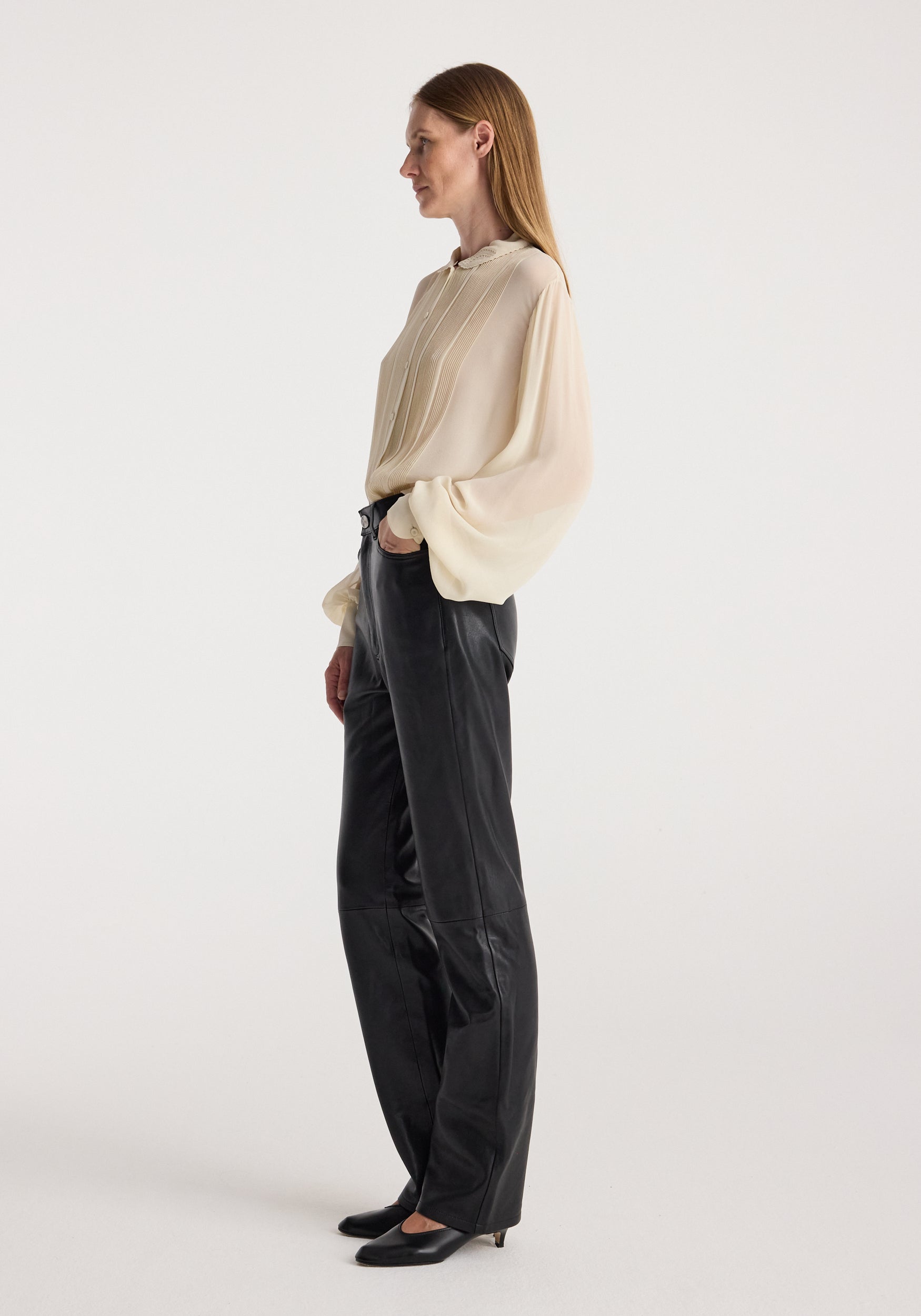 A woman stands in profile wearing a cream-colored, semi-sheer long-sleeve blouse and black wide-leg trousers against a plain light gray studio background.