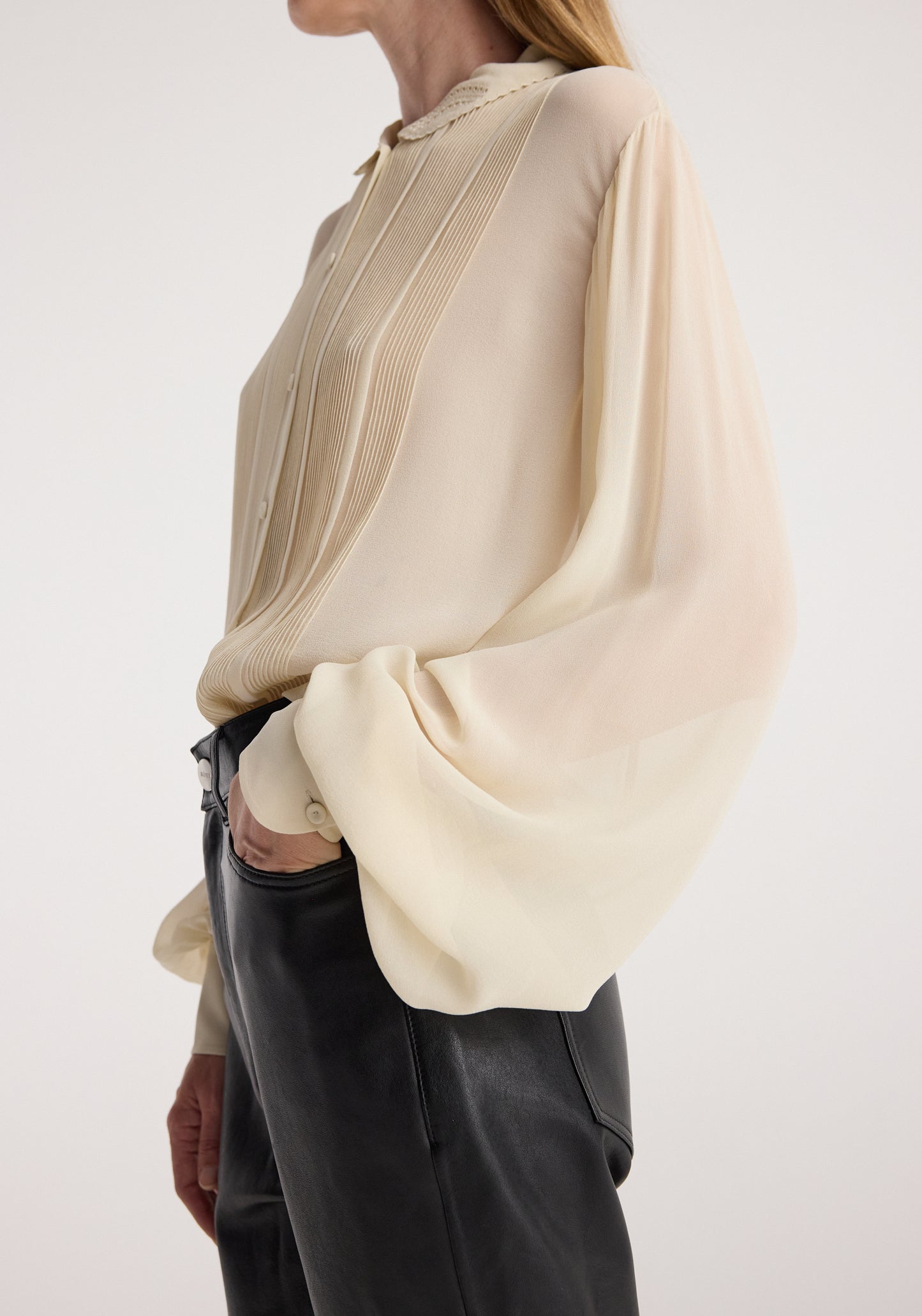 A woman wearing a cream-colored long-sleeve blouse with pleated detailing and black leather pants, shown from a side profile against a plain light background.