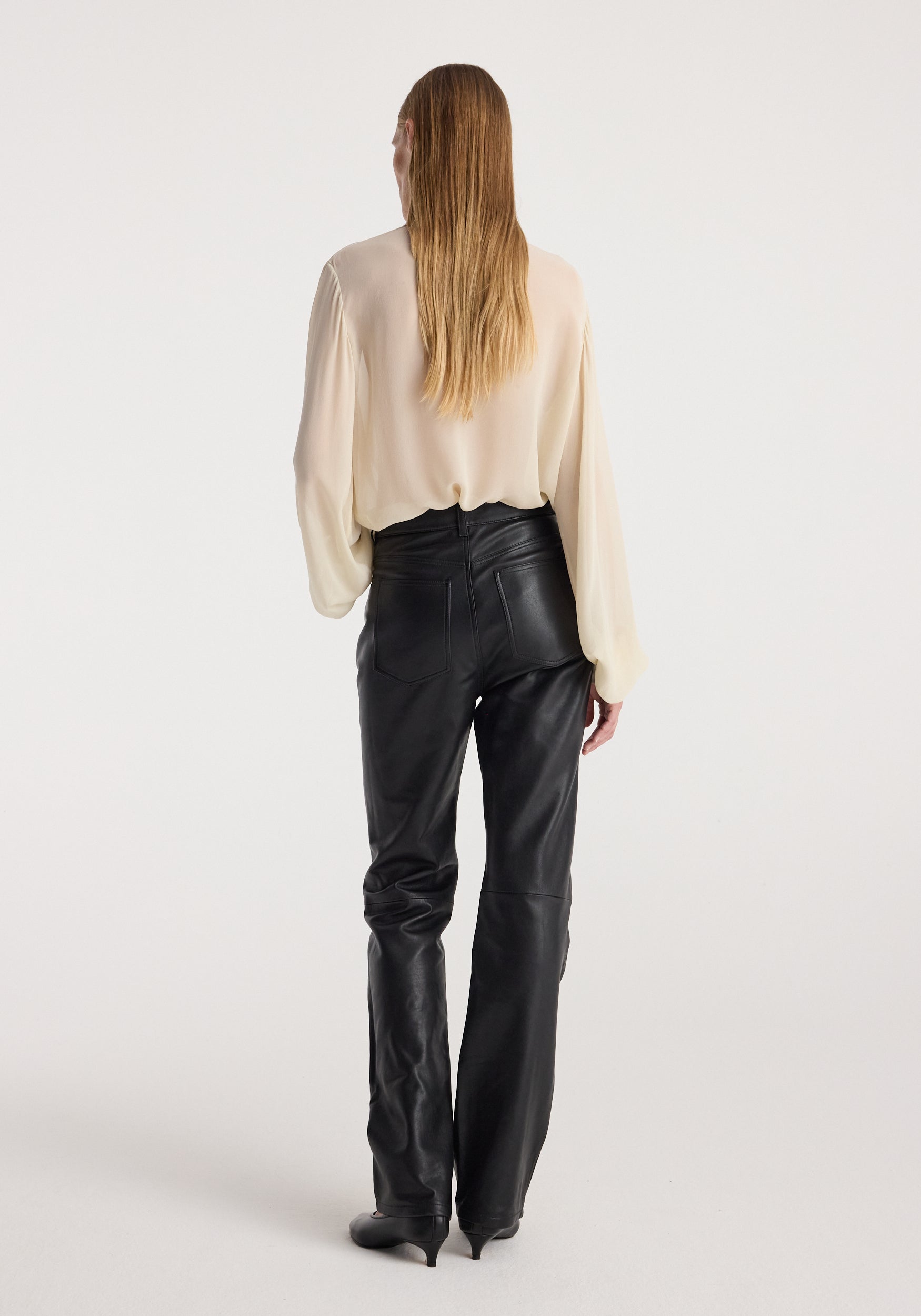 A woman viewed from behind wearing a cream-colored long-sleeve blouse and black leather pants against a plain light gray studio background.