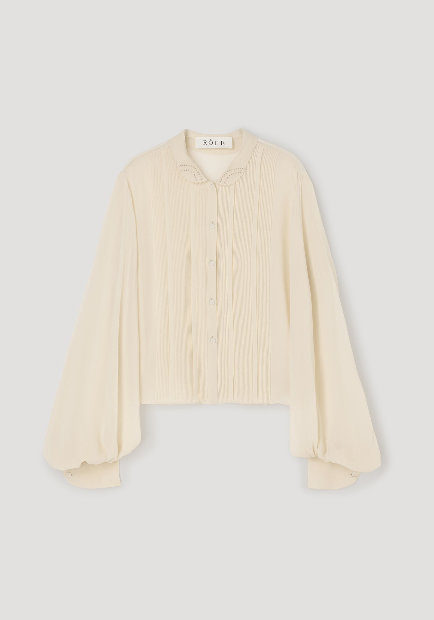 A cream-colored long-sleeve blouse with pleated detailing on the front and a pointed collar, shown flat against a light gray background.
