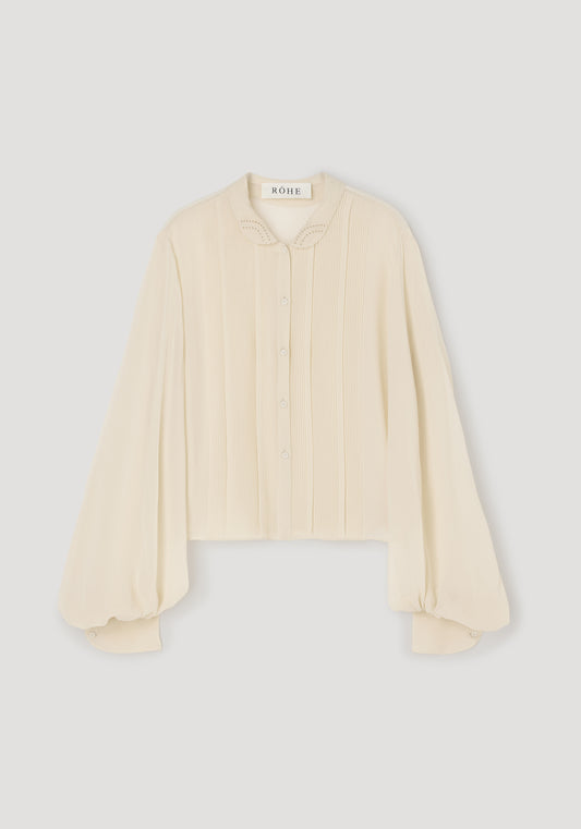 A cream-colored long-sleeve blouse with pleated detailing on the front and a pointed collar, shown flat against a light gray background.