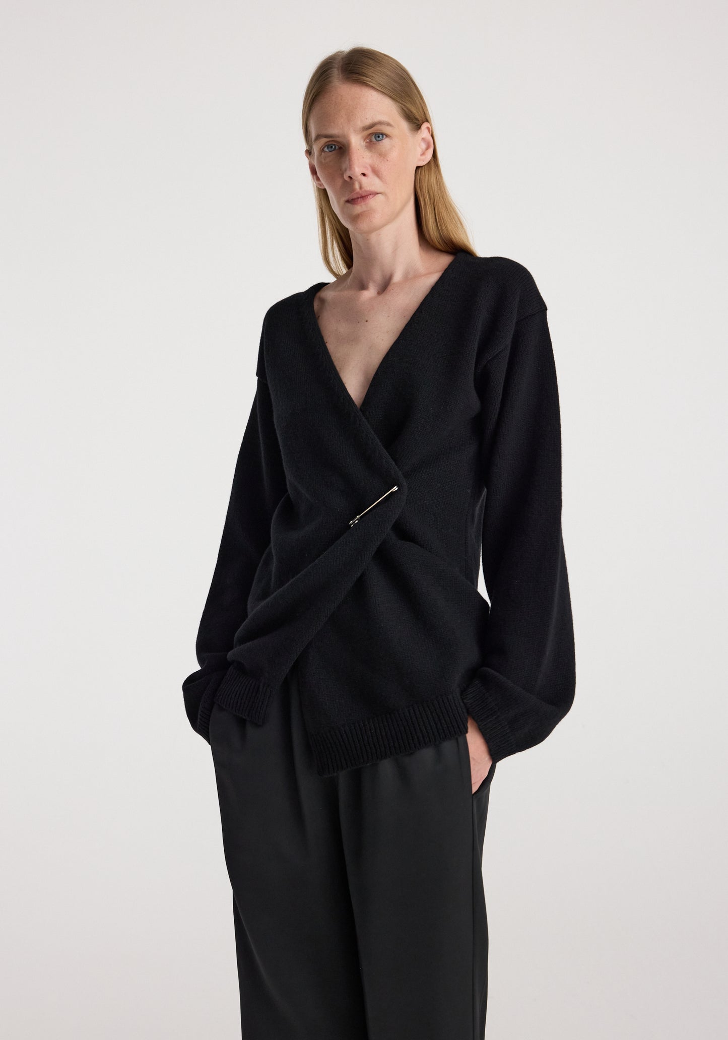 A woman modeling a black knit wrap-style cardigan with a deep V-neck, long sleeves, and a twisted front detail, paired with black wide-leg trousers against a plain light gray studio background.