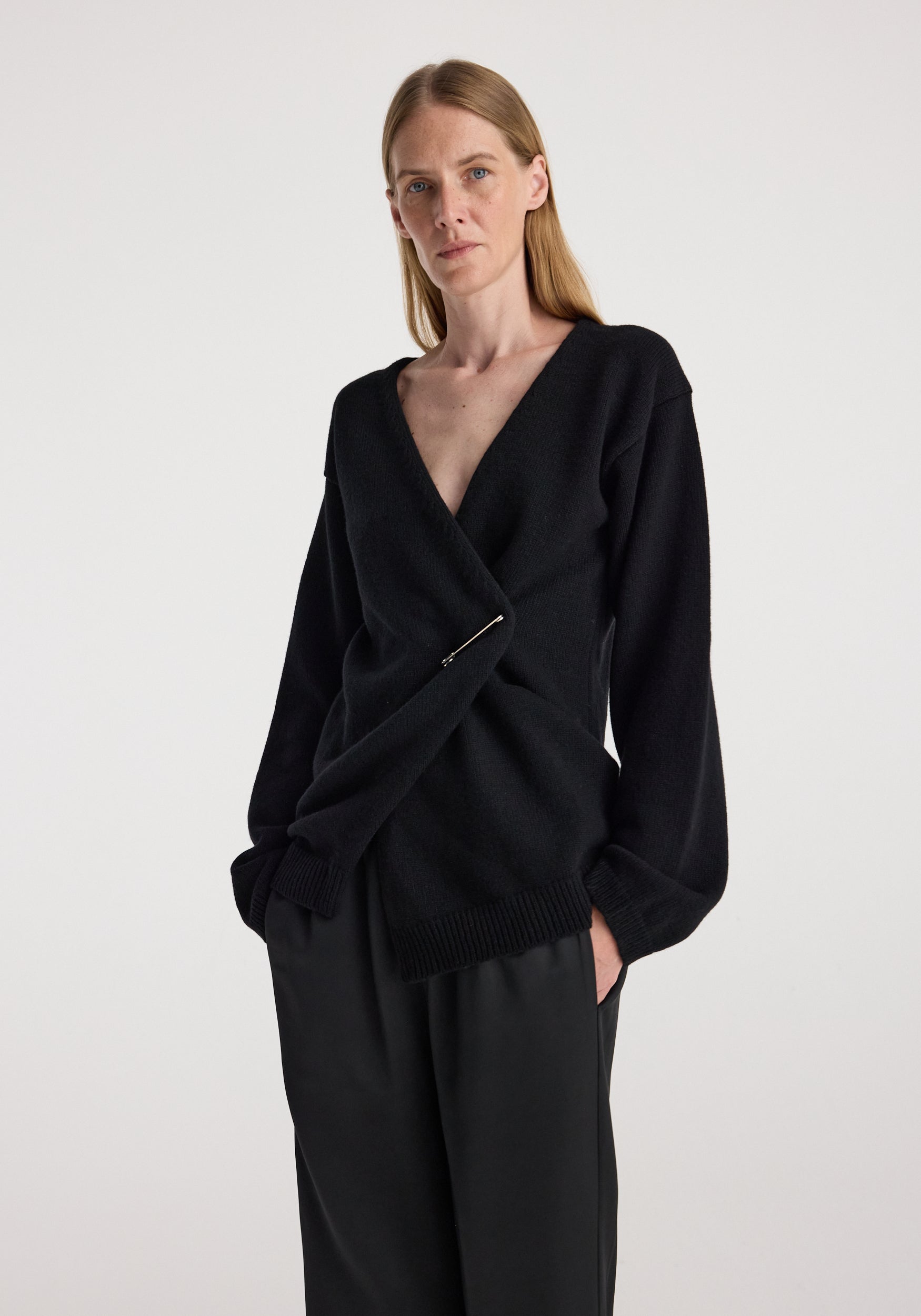 A woman modeling a black knit wrap-style cardigan with a deep V-neck, long sleeves, and a twisted front detail, paired with black wide-leg trousers against a plain light gray studio background.