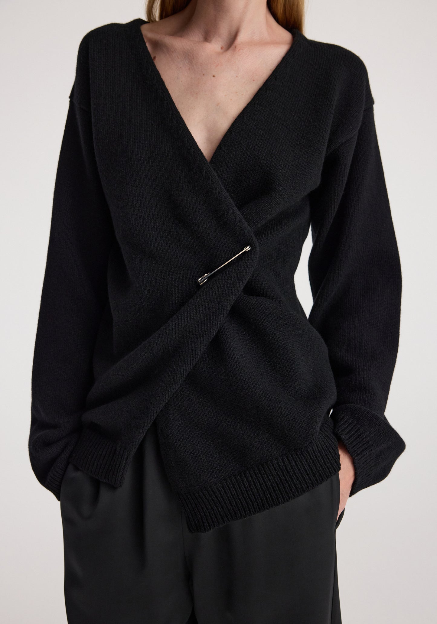 A close-up of a model wearing a black knit wrap sweater with a safety pin detail at the front, paired with black trousers against a light gray background.
