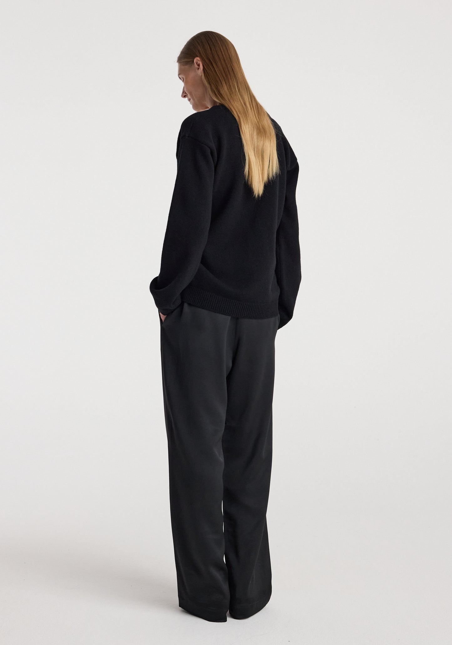 A woman with long blonde hair is shown from the back wearing a black knit sweater and matching black wide-leg trousers against a plain light gray studio background.