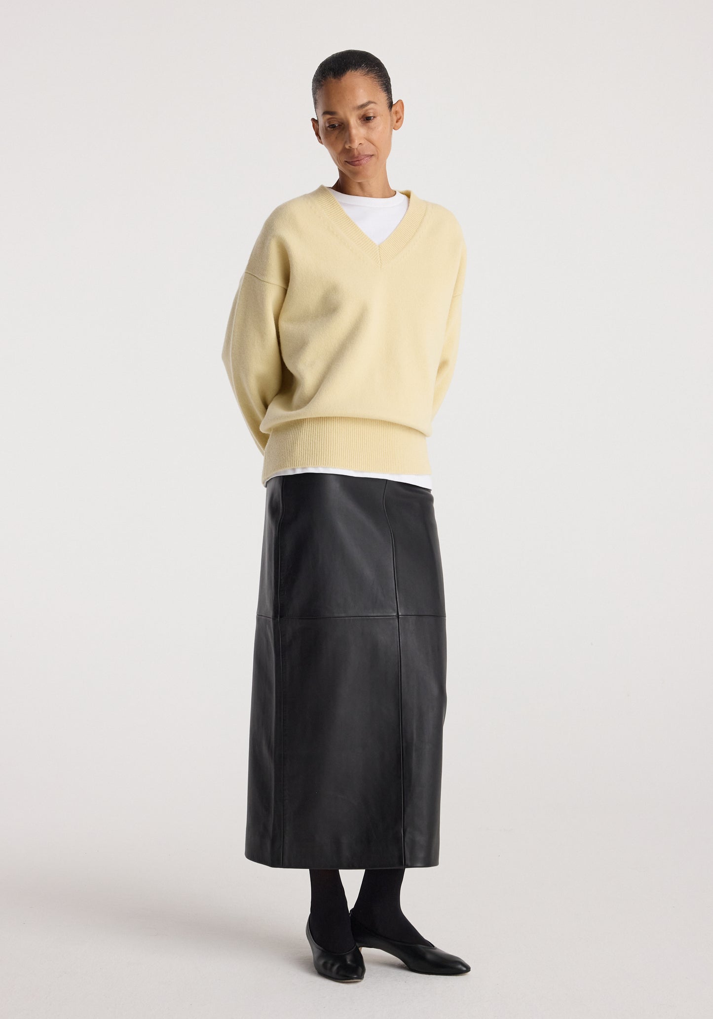 A model stands against a light gray studio background wearing a pale yellow V-neck knit sweater over a white undershirt, a long black leather-look midi skirt, black socks, and black pointed shoes.