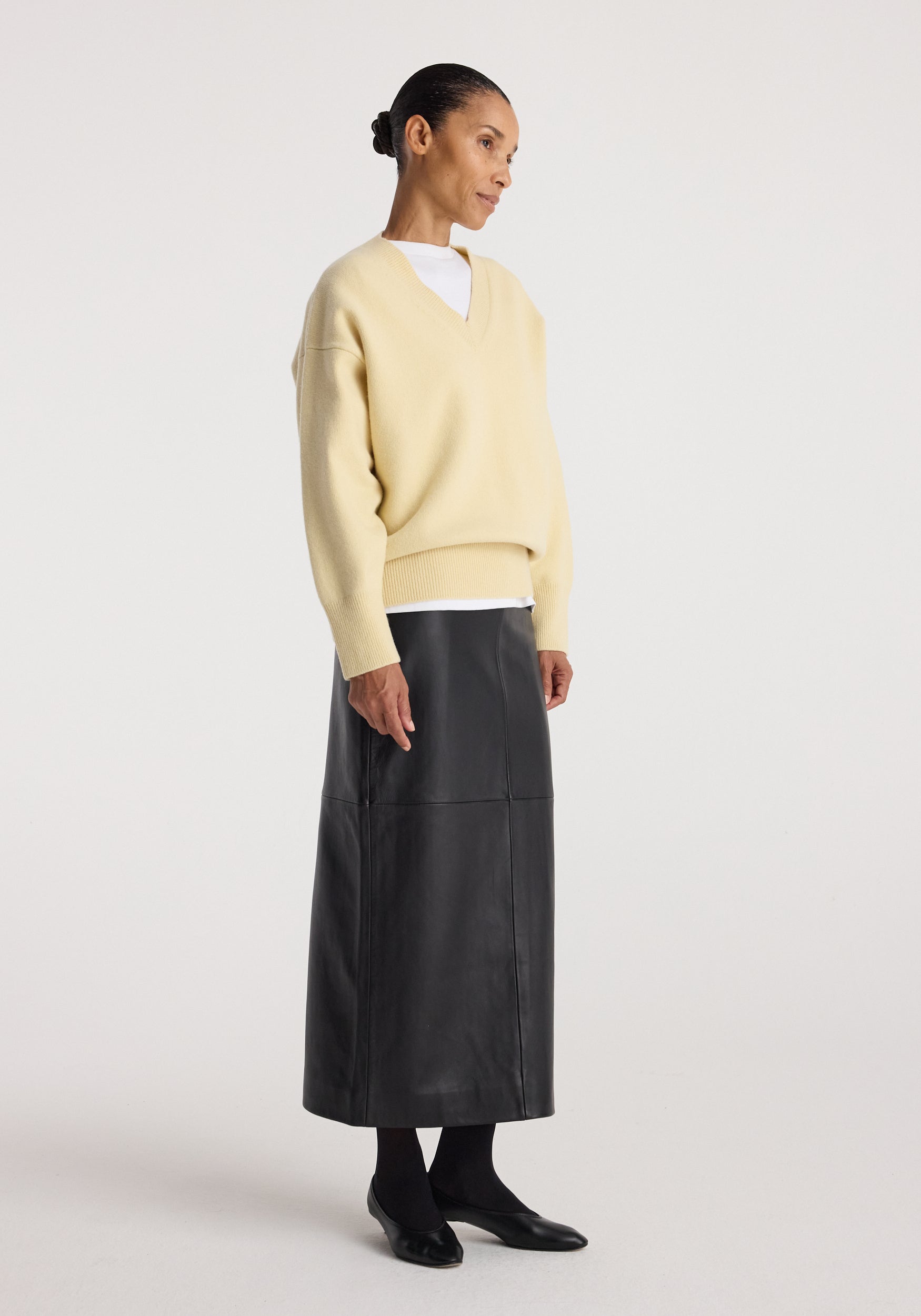 A model is shown in profile wearing a pale yellow knit sweater over a white crewneck top, paired with a long black leather skirt and black shoes, against a plain light gray studio background.