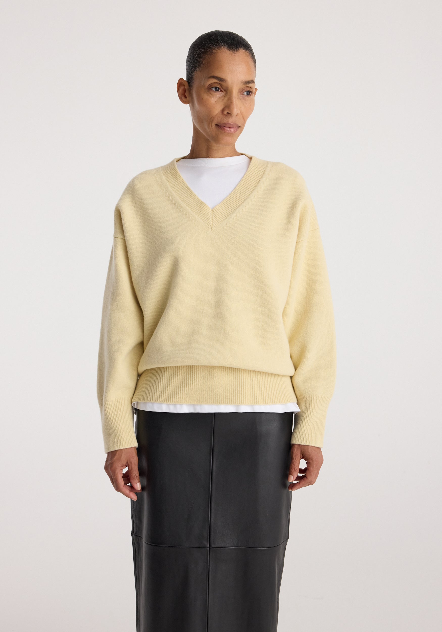 A model wearing a pale yellow V-neck knit sweater over a white undershirt and a black leather skirt, photographed against a plain light gray studio background.