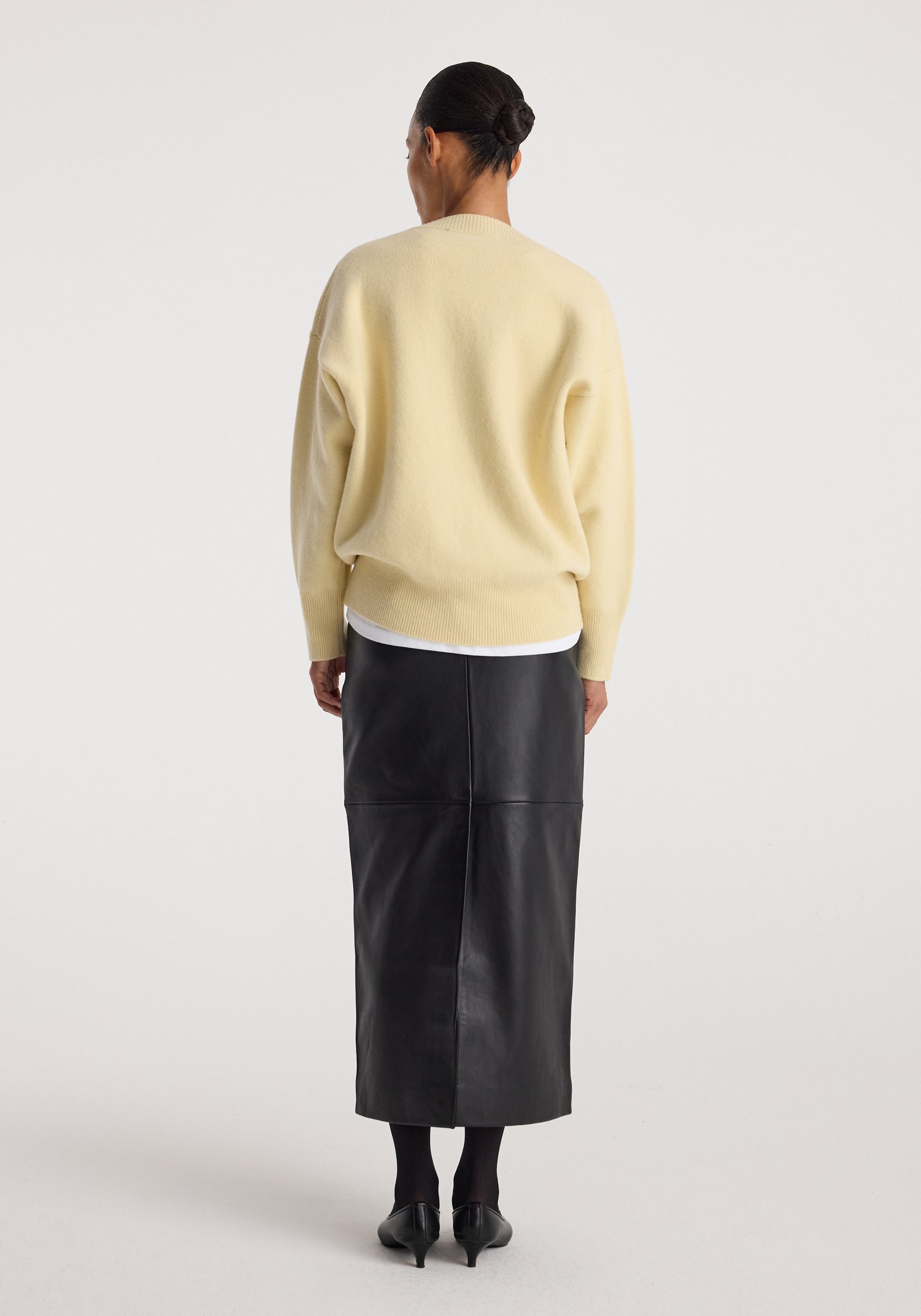 A model shown from the back wearing a pale yellow knit sweater, a black leather midi skirt, black tights, and black heels, against a plain light gray studio background.