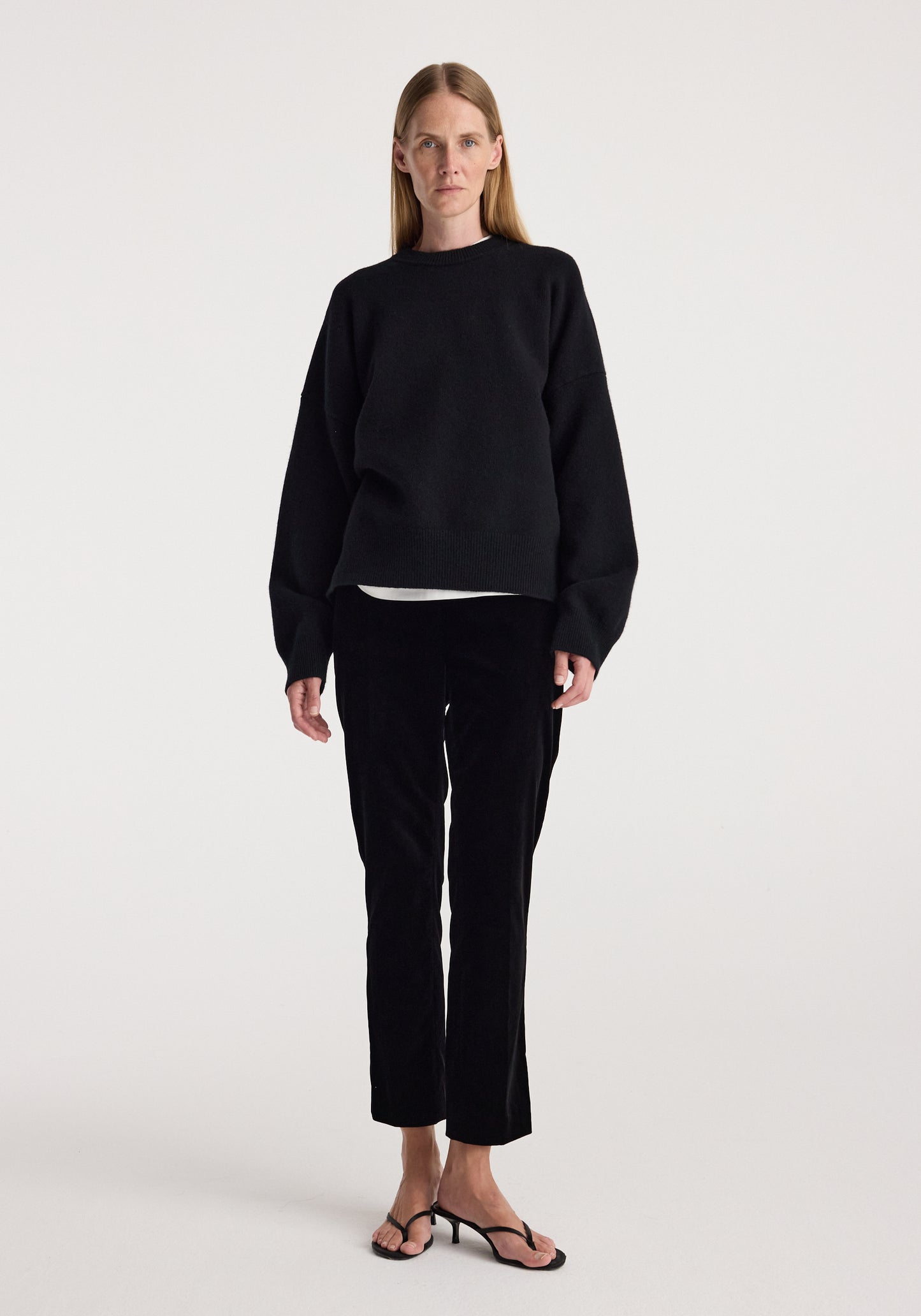 A woman stands against a plain light gray studio background wearing a black long-sleeve top, black cropped pants, and black heeled sandals.