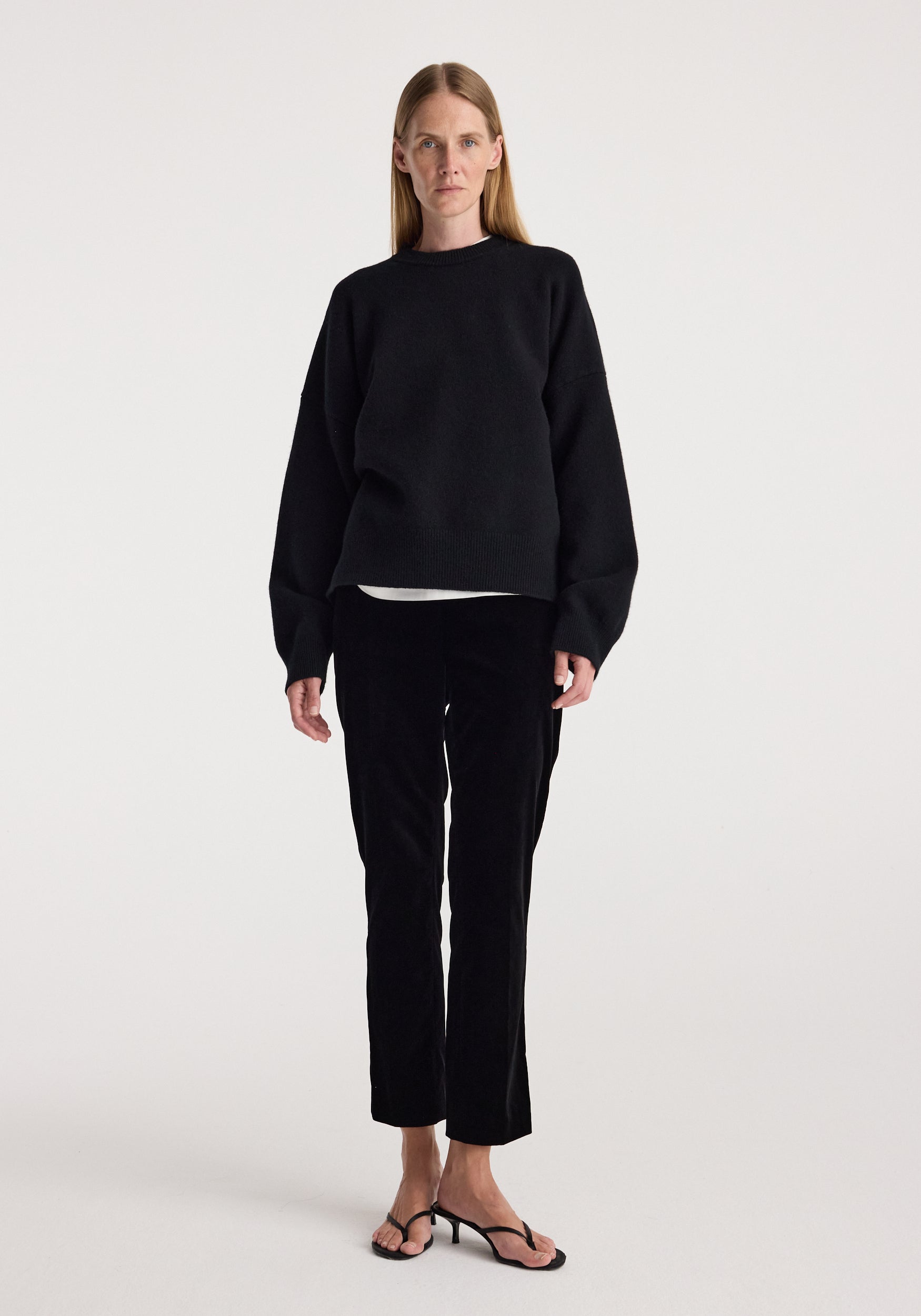 A woman stands against a plain light gray studio background wearing a black long-sleeve top, black cropped pants, and black heeled sandals.