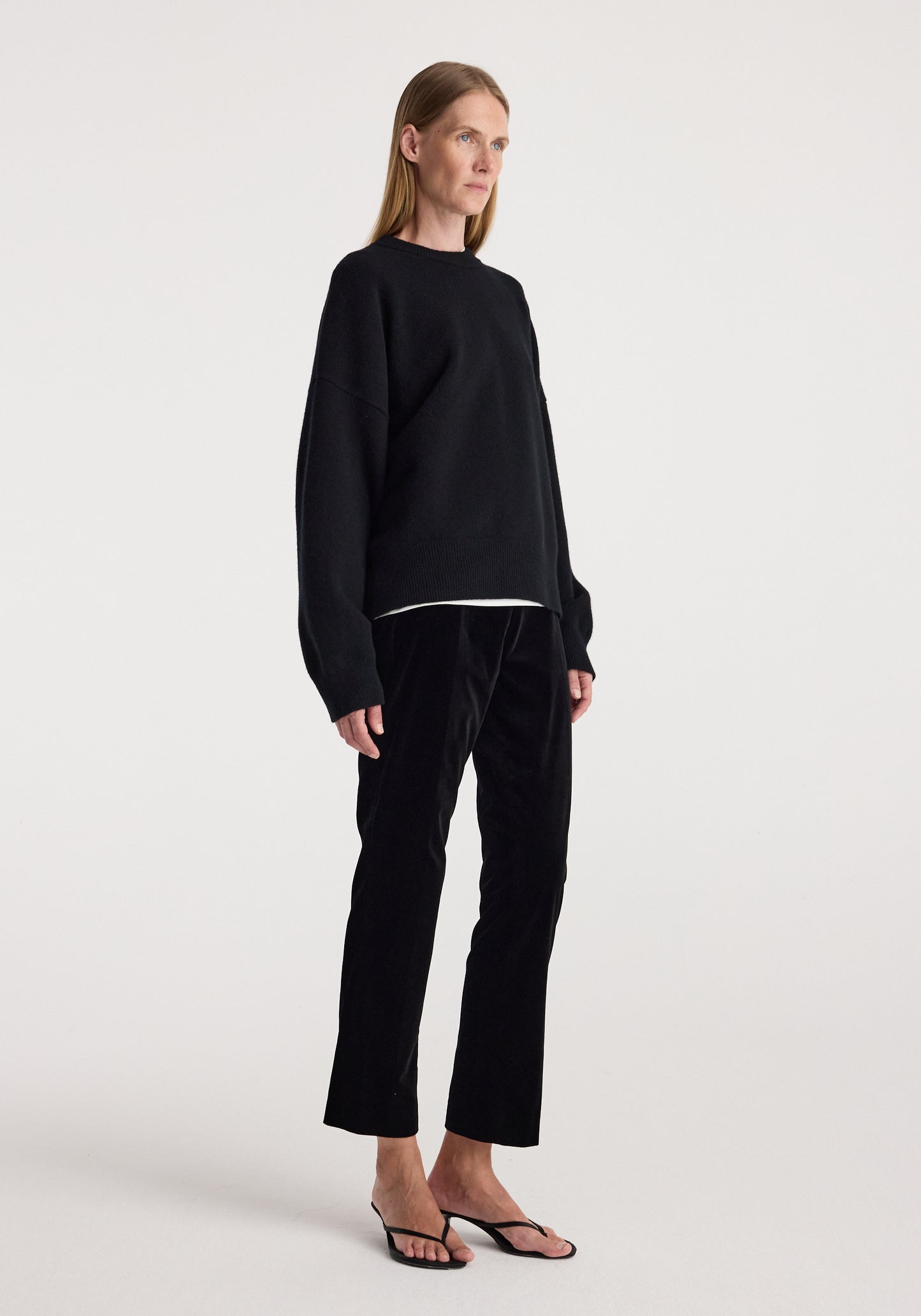 A model stands in profile wearing a black long-sleeve crewneck sweater, black cropped trousers, and black flat sandals against a plain light gray studio background.