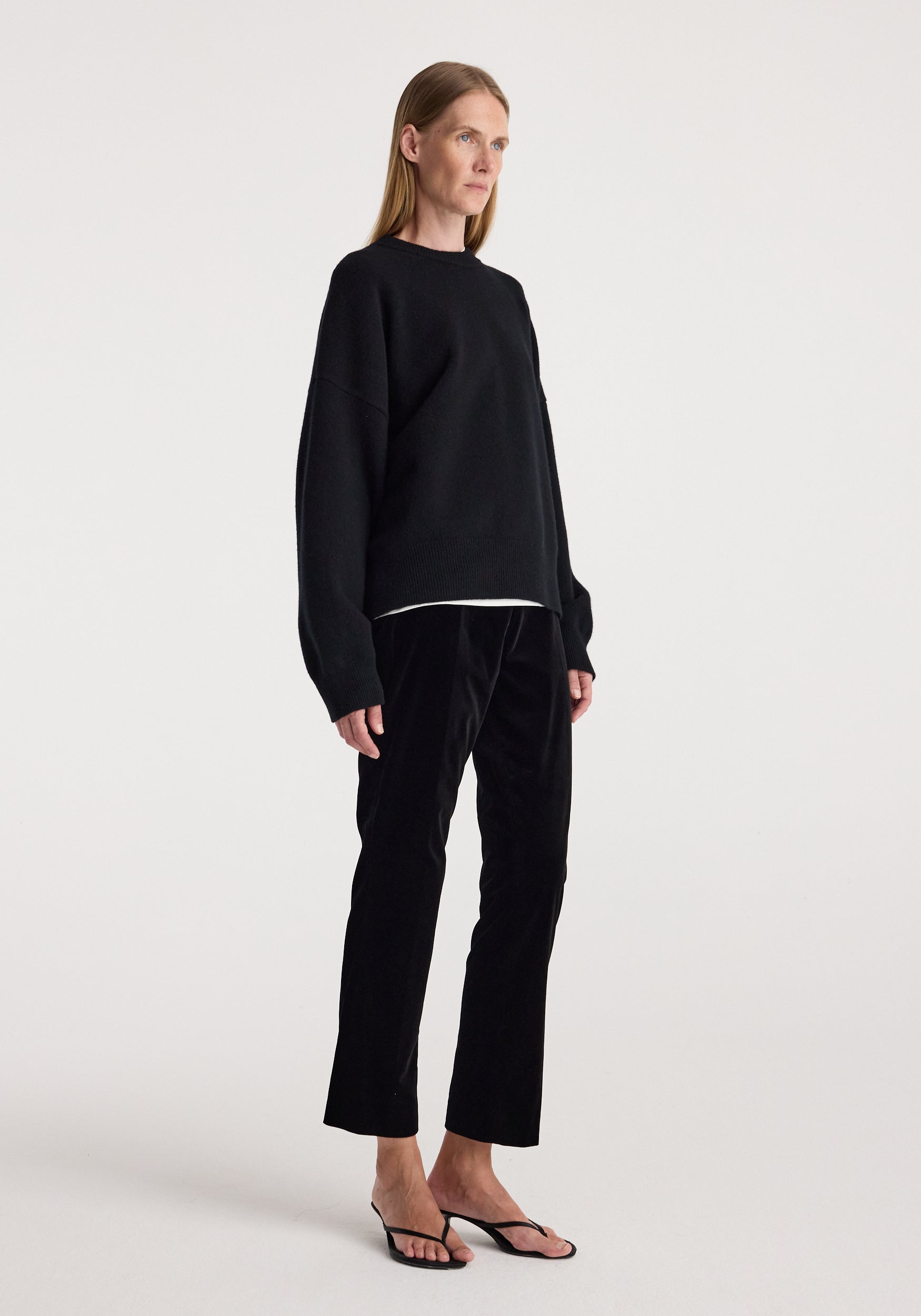 A model stands in profile wearing a black long-sleeve crewneck sweater, black cropped trousers, and black flat sandals against a plain light gray studio background.