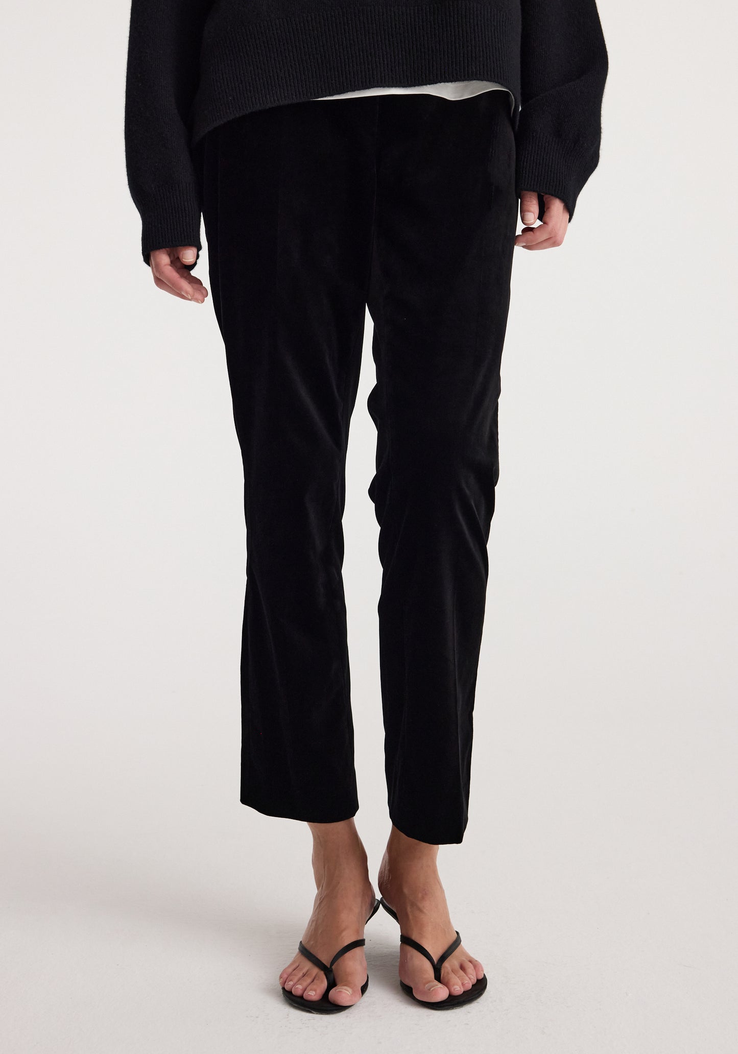 Black cropped trousers worn by a model, paired with a black sweater and black thong sandals, photographed against a clean white studio background.
